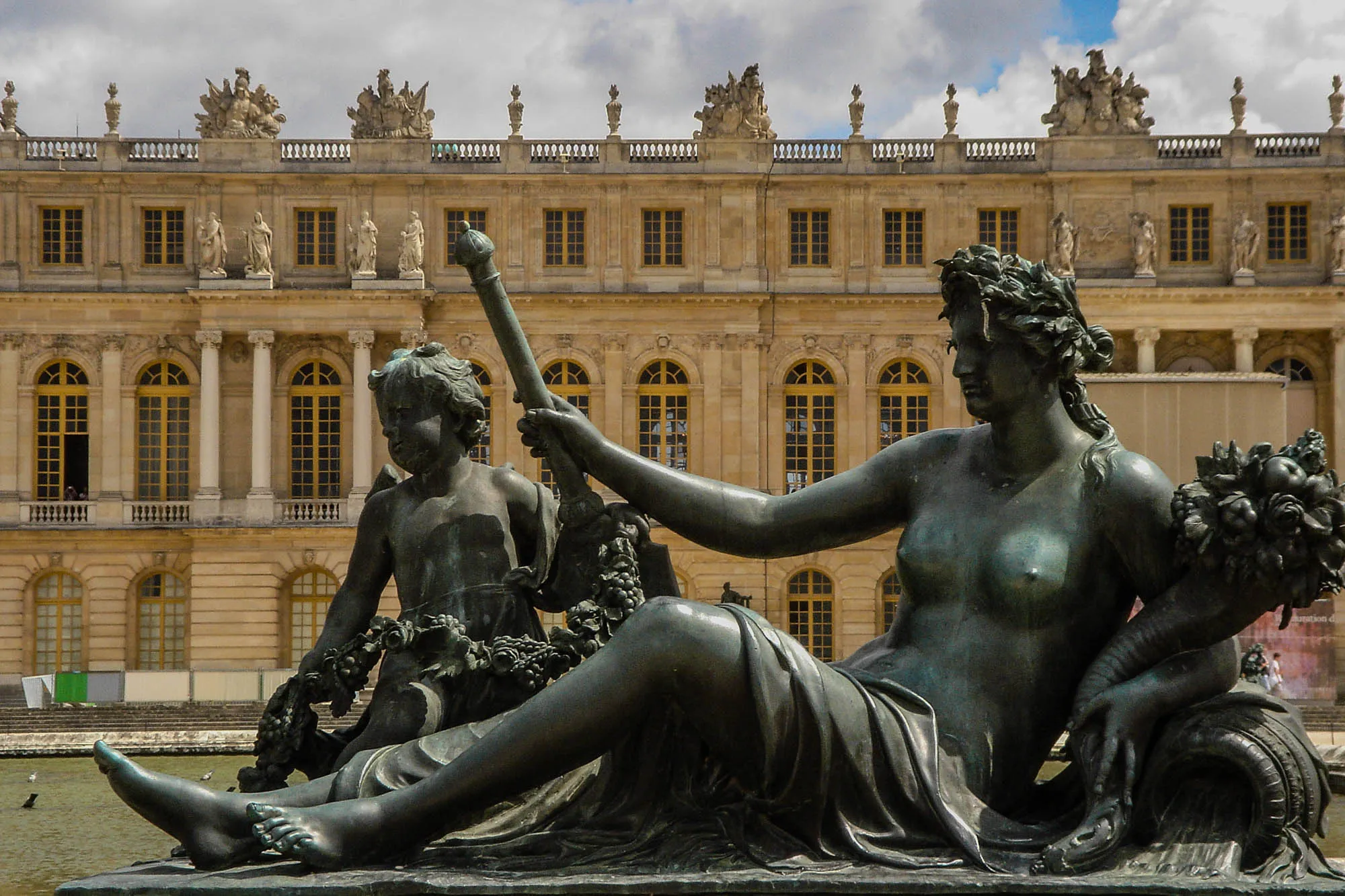 Bronze statue of a nude woman with leaves in her hair and a scepter in her right hand, reclining, held up by her left elbow on a potm a bouquet of flowers cradled in her left arm. Water flows out of the pot. A putto stands behind her 