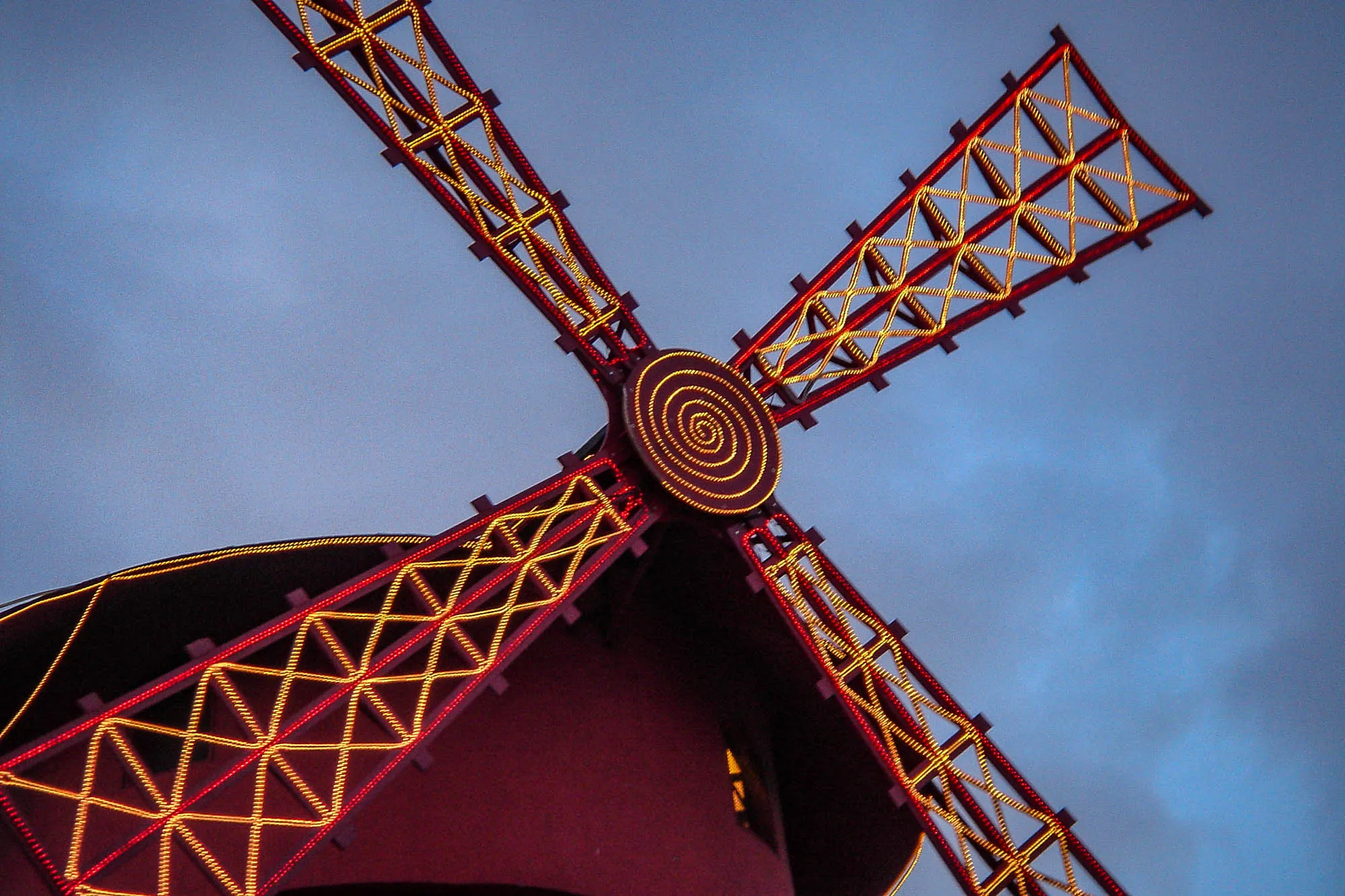 The top of a red windmill, its blades covered in criss-cross warm white lighting, bordered by red lighting. The center of the blades has a spiral of warm white lighting.