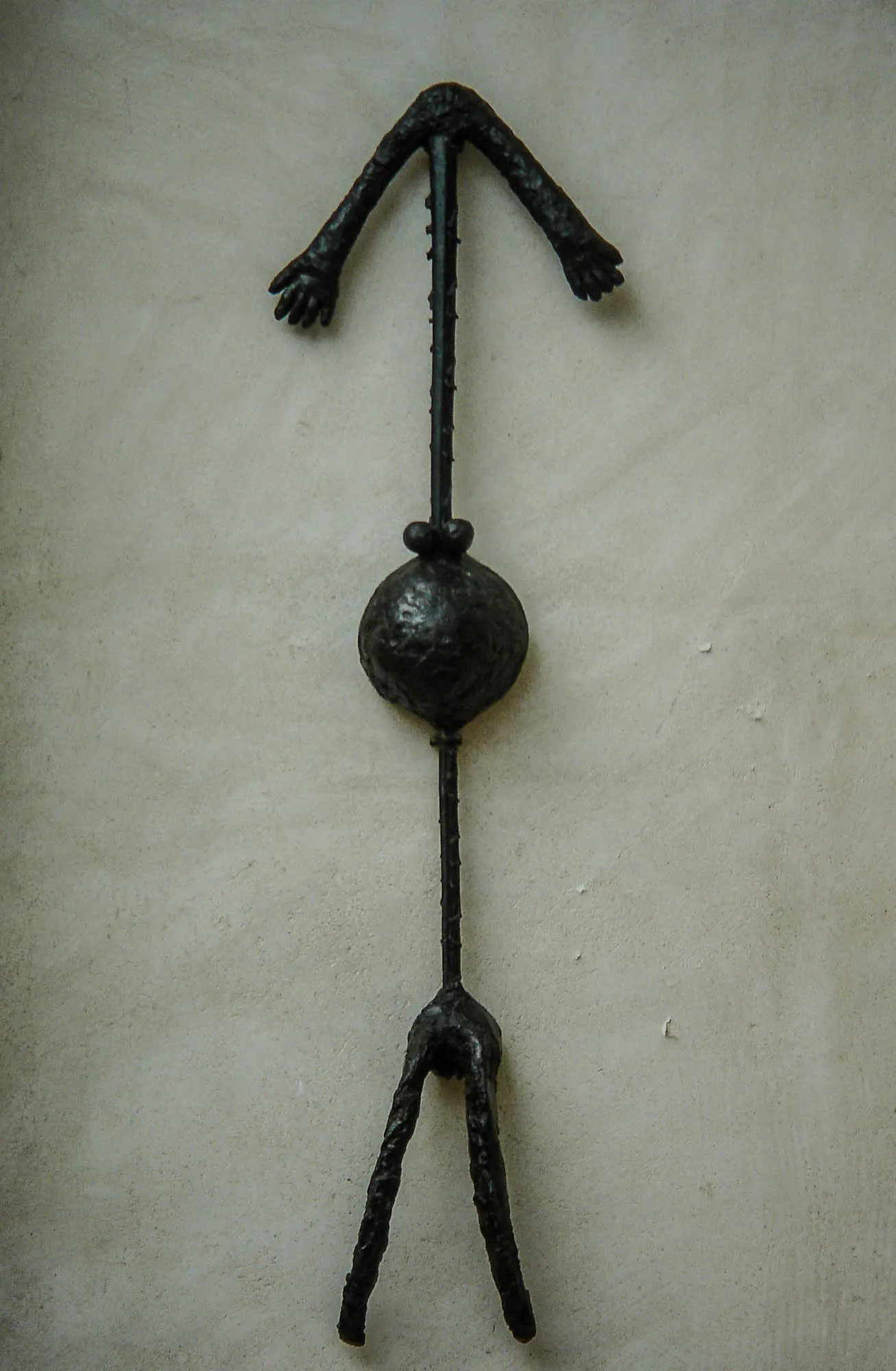 A black metal sculpture of an arrow, pointing up, against a cream stucco wall. The arrow head is mead of two arms with small hands at the end. The middle has a ball, and the fletching looks like two legs, giving the arrow an overall appearance resembling a person.