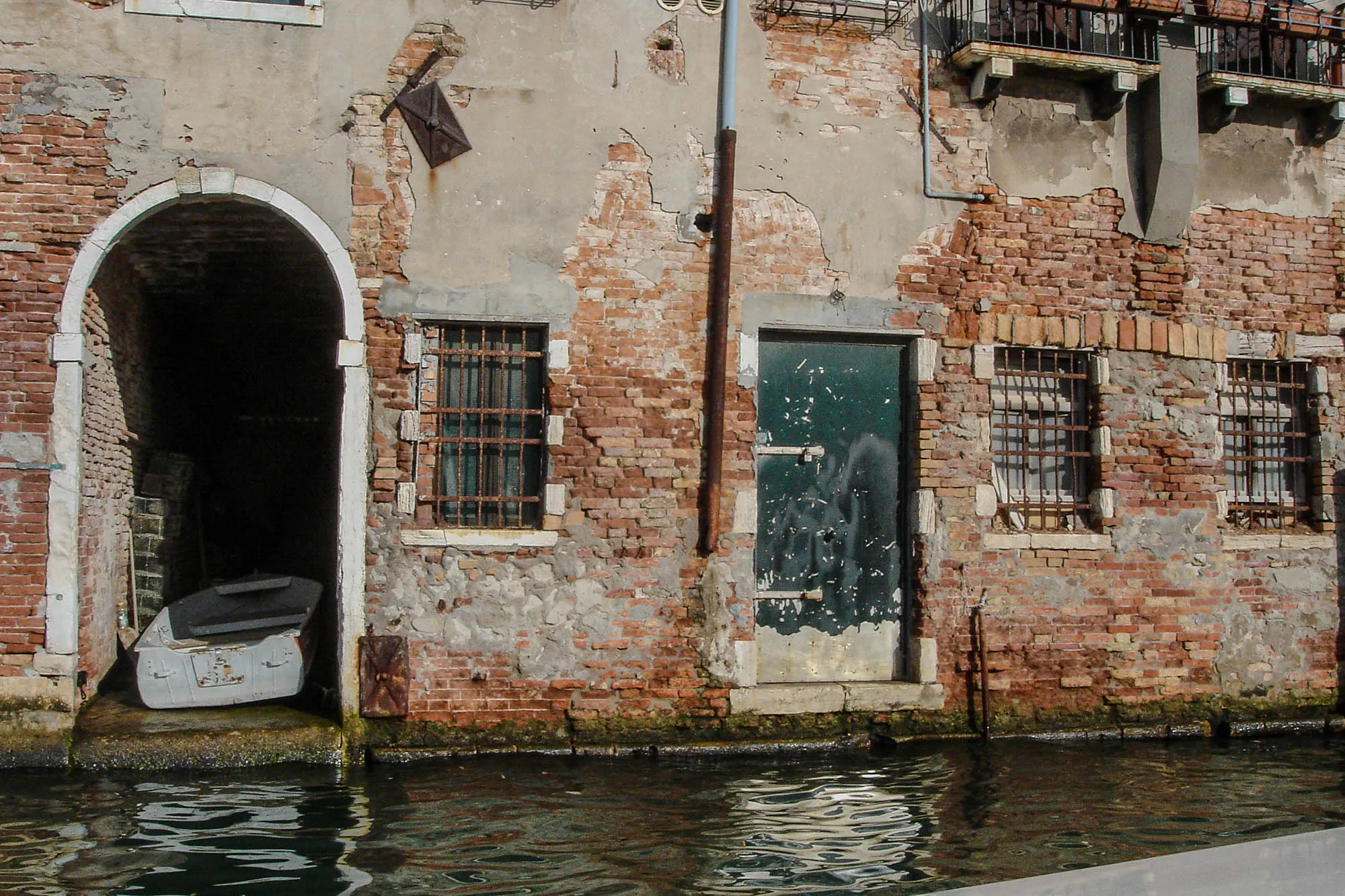 An arch cutout next to a blue metal door of a brick building with faded stucco covering. A small wite well-worn boat is in it. The building has windows with rusted rebarb covers on it, and is on the river in Venice.