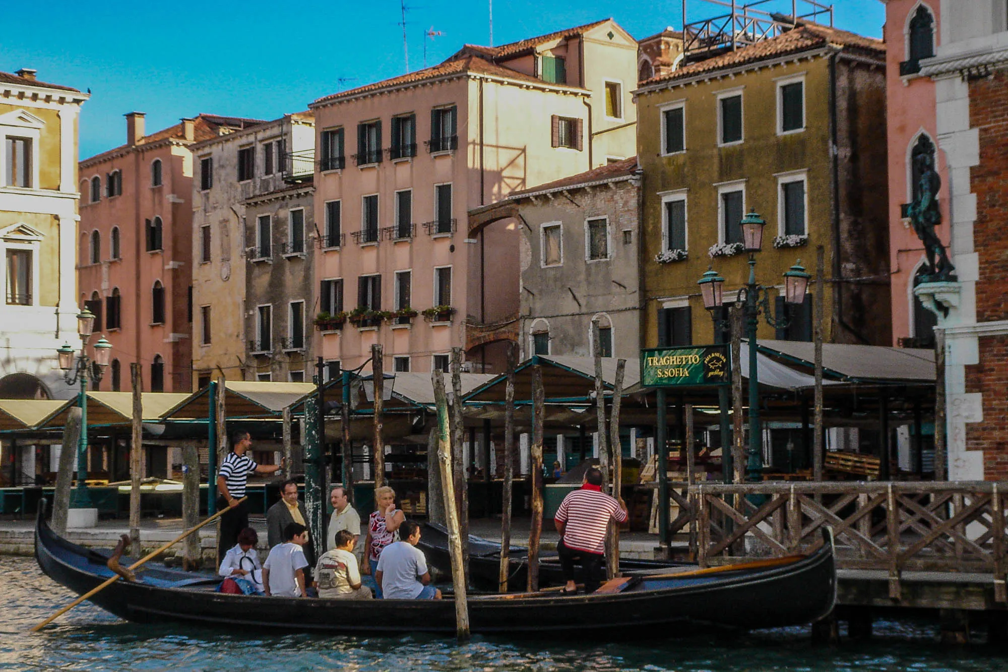 A gondola in Venice with 8 passengers and 2 gondoliers parked Pastel colored buildings, no more than 5 stories tall, line the doc the gondola is parked at.