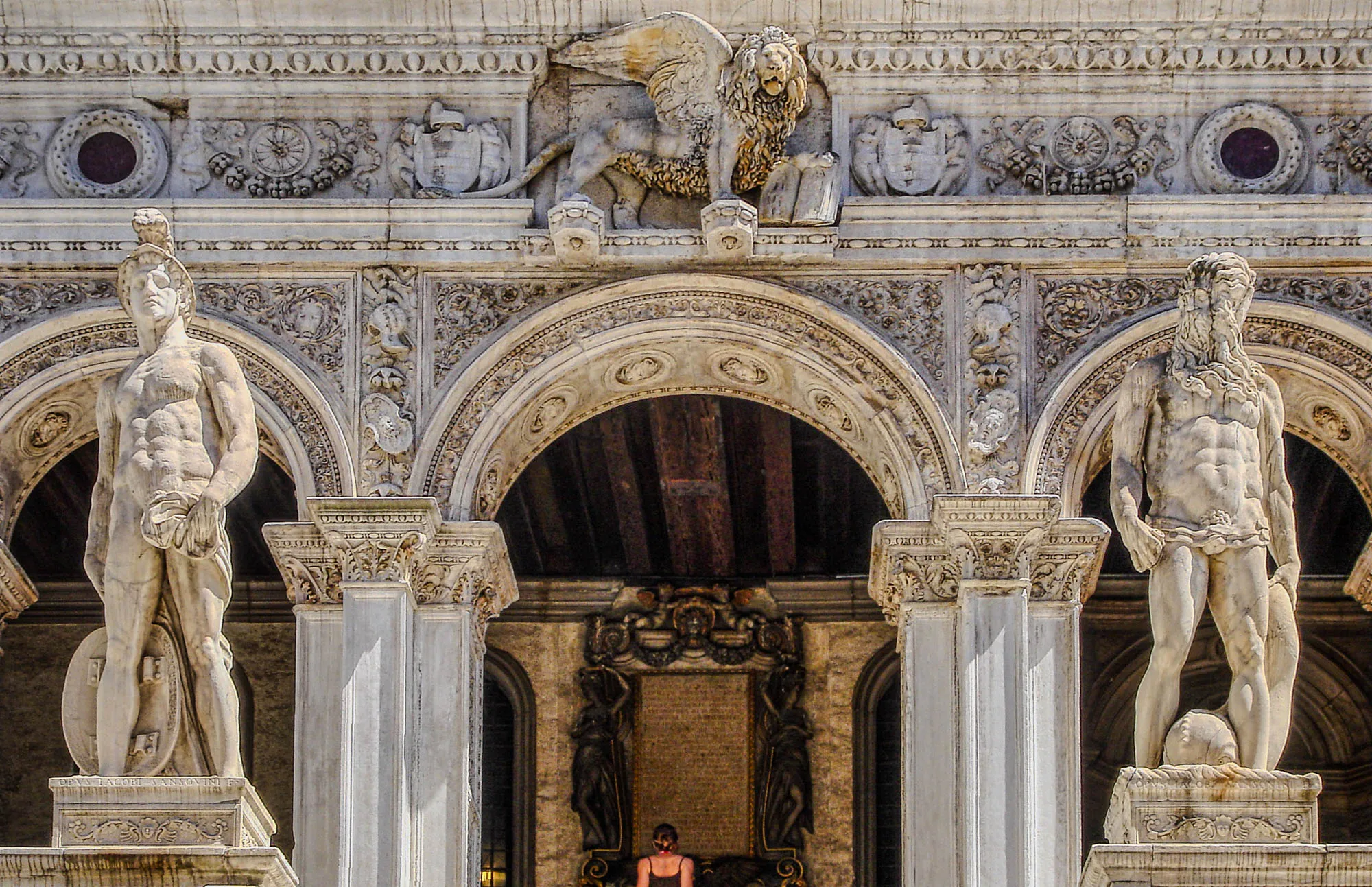 Two large marble statues of nude men, the one on the left depicting Mars with a shield behind his leg and a soldier's helmet on, the one on the righte depecting Neptune carying a large sea creature. Behind them, ornate archways. A lion with eagle wings and a halo, its left front paw on a book, stands above the center arch. A woman, as large as one of the statue's torsos, leans back and observers the plaque inside under the center arch.