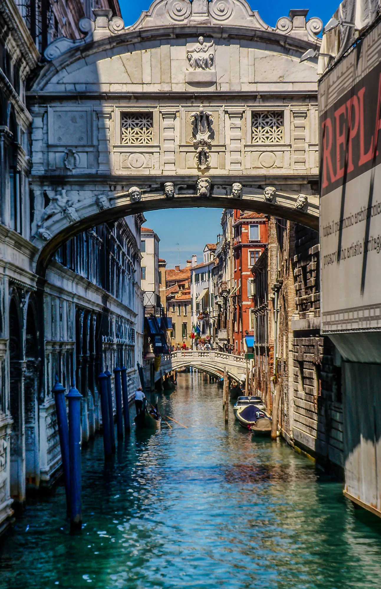 A river street in Venice. A gondola on one side, a parked boat on the other, buildings lining both sides. An archway frames the front of the scene, and a bridge with pedestrians is in the background.