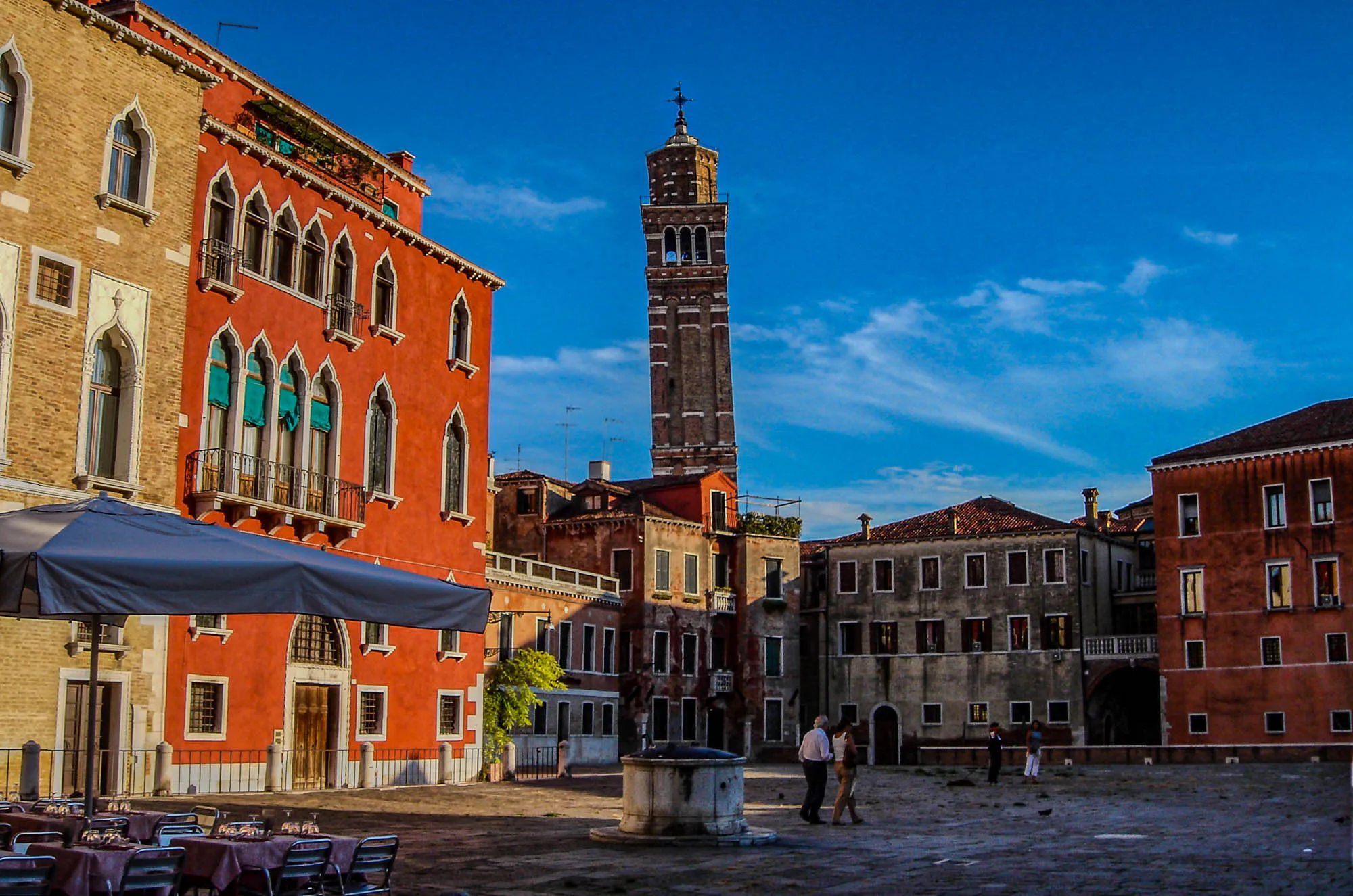 A square in Venice, Italy, with a brick and red building to the left, lower three store buildings forming a corner , and a tall tower jutting up from behind the corner of the buildings. A couple walks through the square.