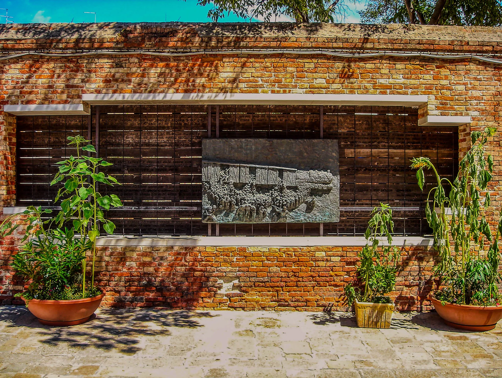 A bronze plaque with embossed artwork depicting a train with box cars and a large number of people being forced into those box cars by armed individuals. It is situated inside a cutout in a brick façade with wooden planks behind it and words etched into the planks. In front of the cutout, not blocking the plaque, are some small potted plants.