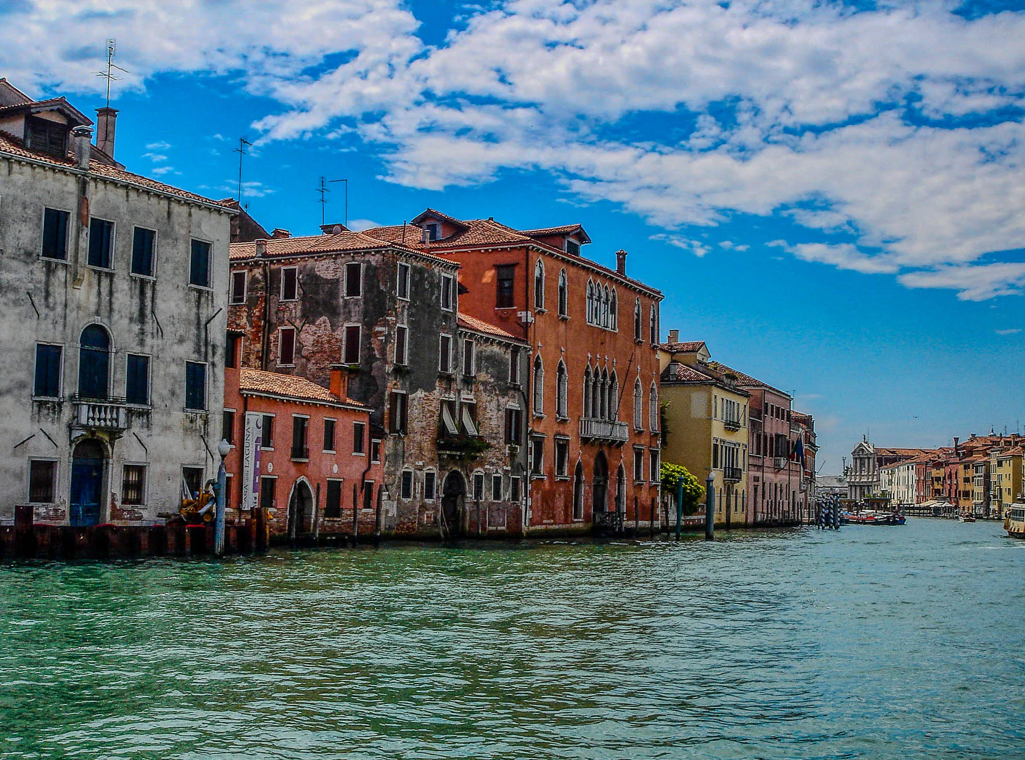 The image shows a view of a canal in Venice, Italy. A row of buildings lines the canal, with a large, bright building on the right. The large building has a lot of windows and is painted orange. The buildings on the left are taller and are painted in a more muted color. The sky is blue with white clouds. The canal is calm and still, and the water is a deep green color. There is a boat in the distance, and a person is standing by a railing on the left side of the image.