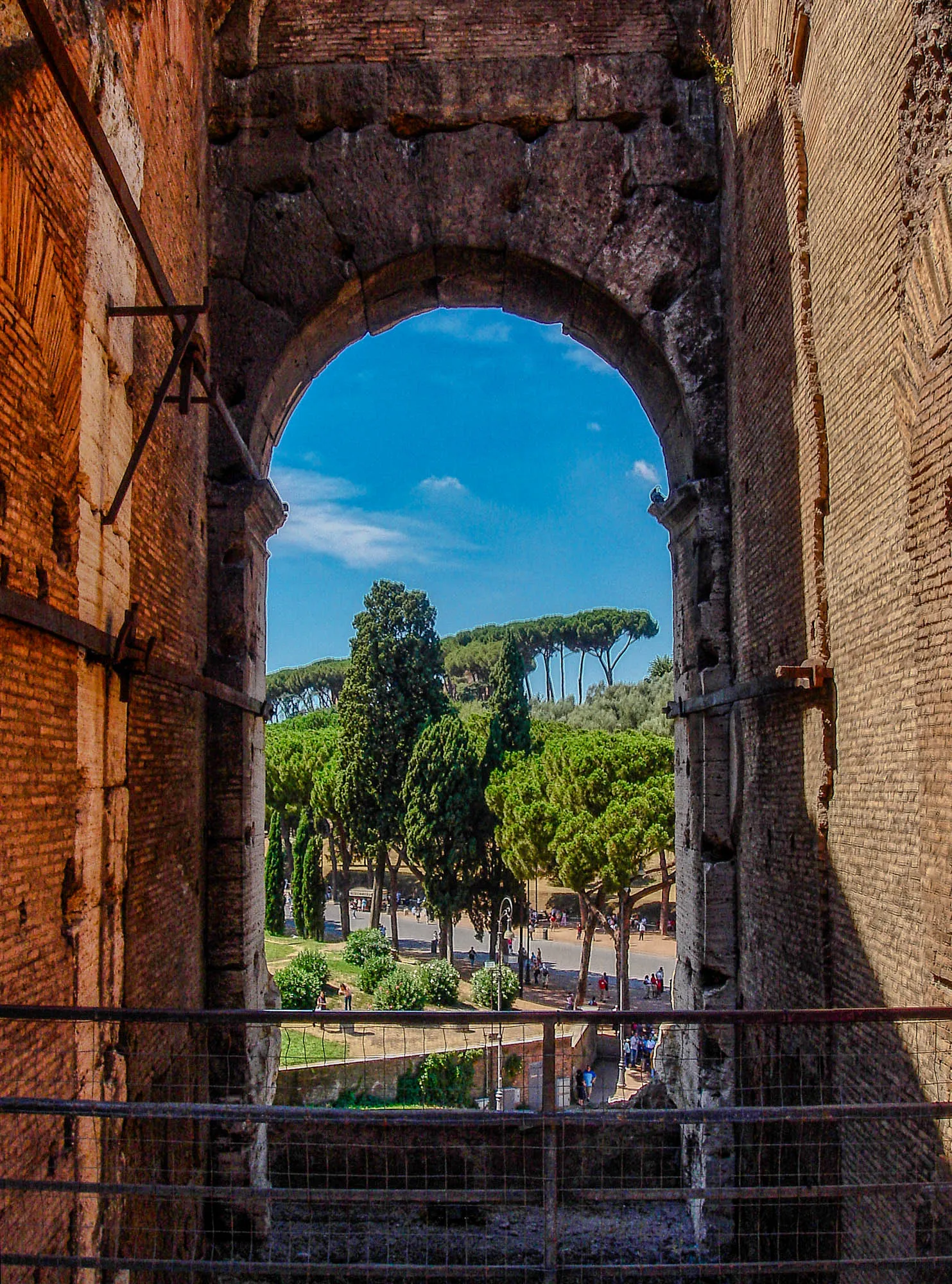 A brick arched window in the Colosseum overlooking a walkway filled with people and lined with trees