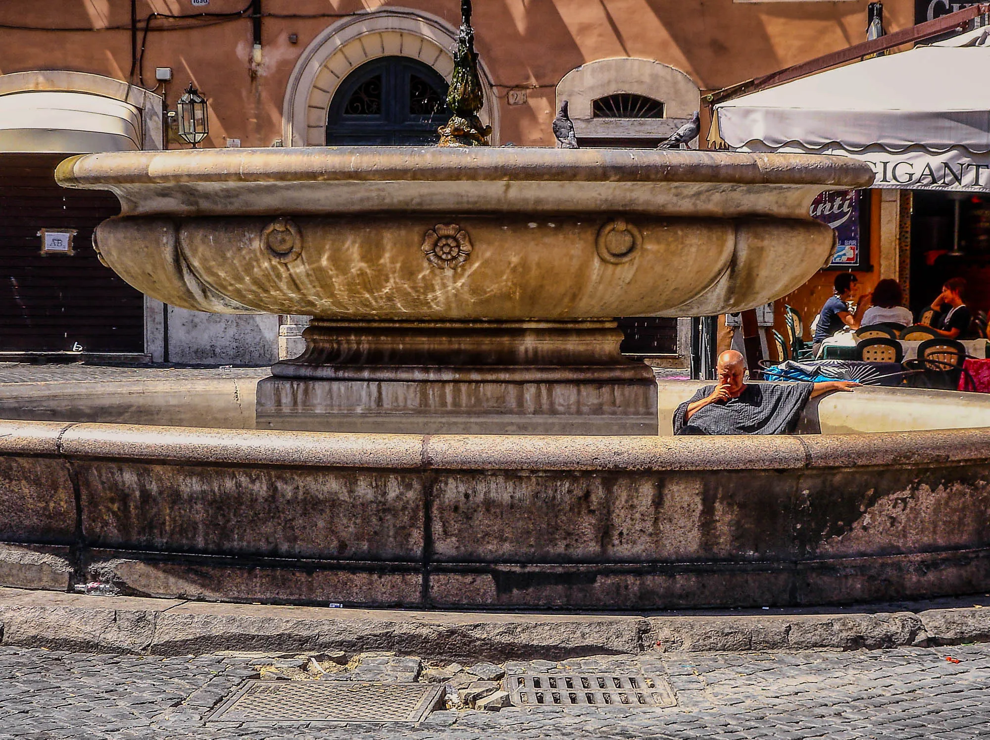 A older man in a grey terrycloth poncho sits inside the lower basin of a two tiwered fountain, resting his hand agains the side, his hand on his chin, deep in contemplation. Two pidgeons take a bath in the upper basin. A group of three women sit at a café in the background, unbothered by this man.