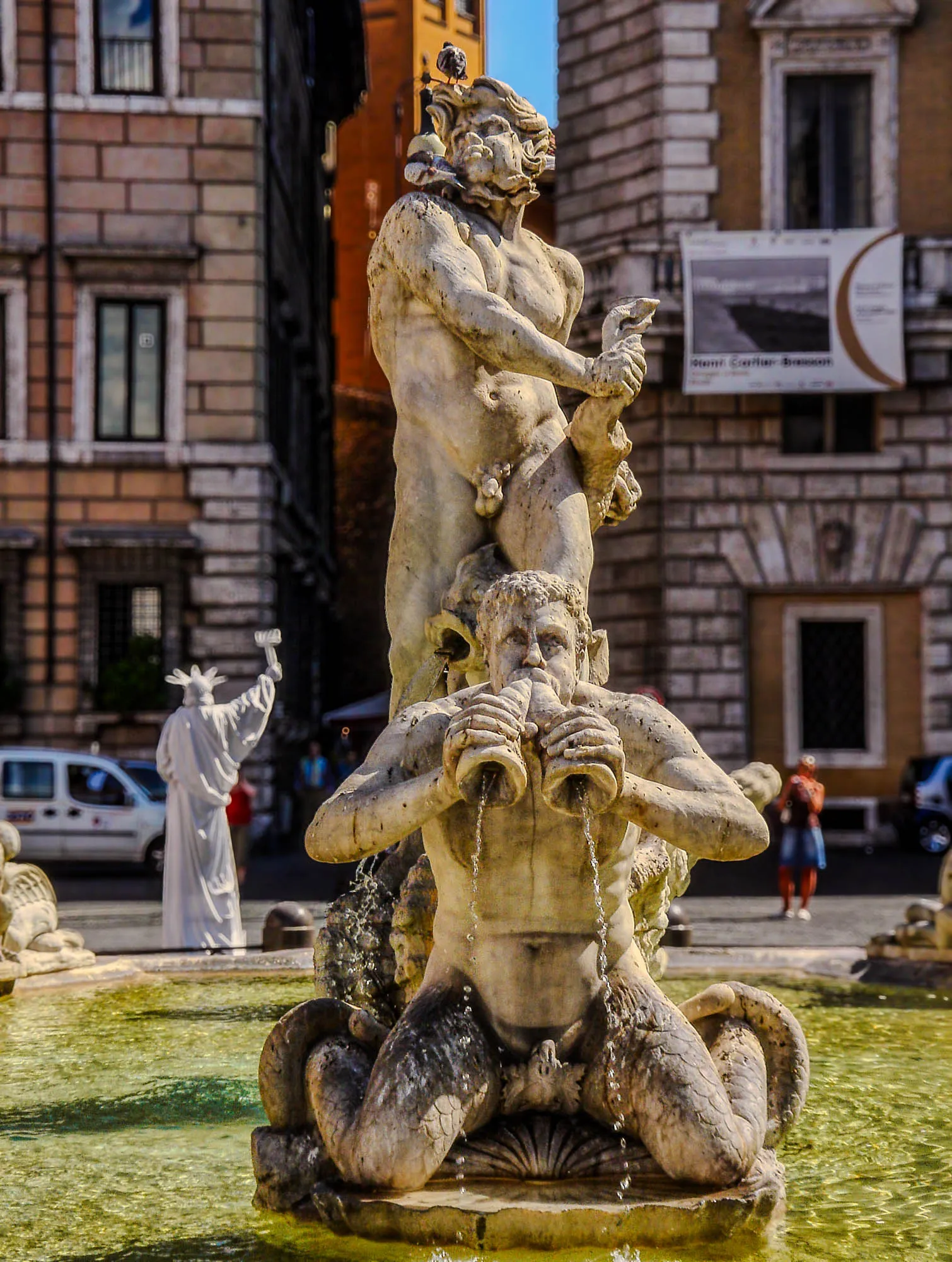 A marble statue in the middle of a fountain basin of a nude man standing upright, his right arm crosed over his body, holding the tail of a sea creature, its head between its legs. a Triton, upper half man, lower half scaly legs, kneels in front of him, blowing two shells. Both the fish head and the two shells are expelling water. A white "human statue" of the statue of liberty is behind them.