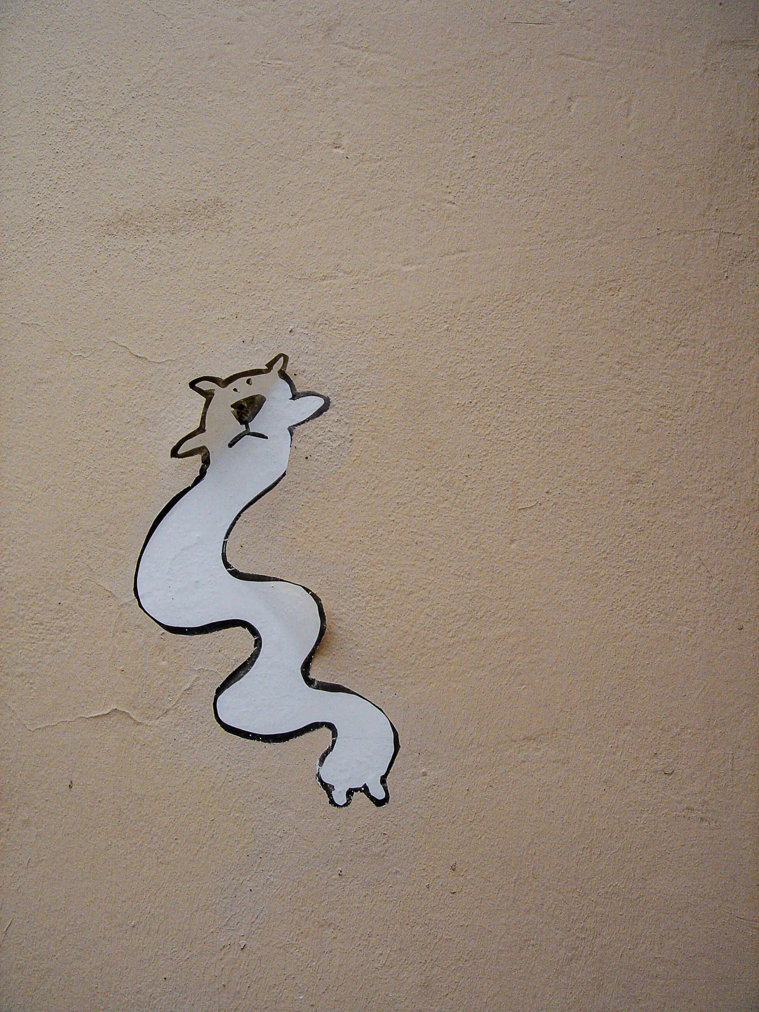 Graffiti of a black-outline on white paper cartoon animal, with a dog like face and long, squiggly body, pasted onto a tan stucco wall.