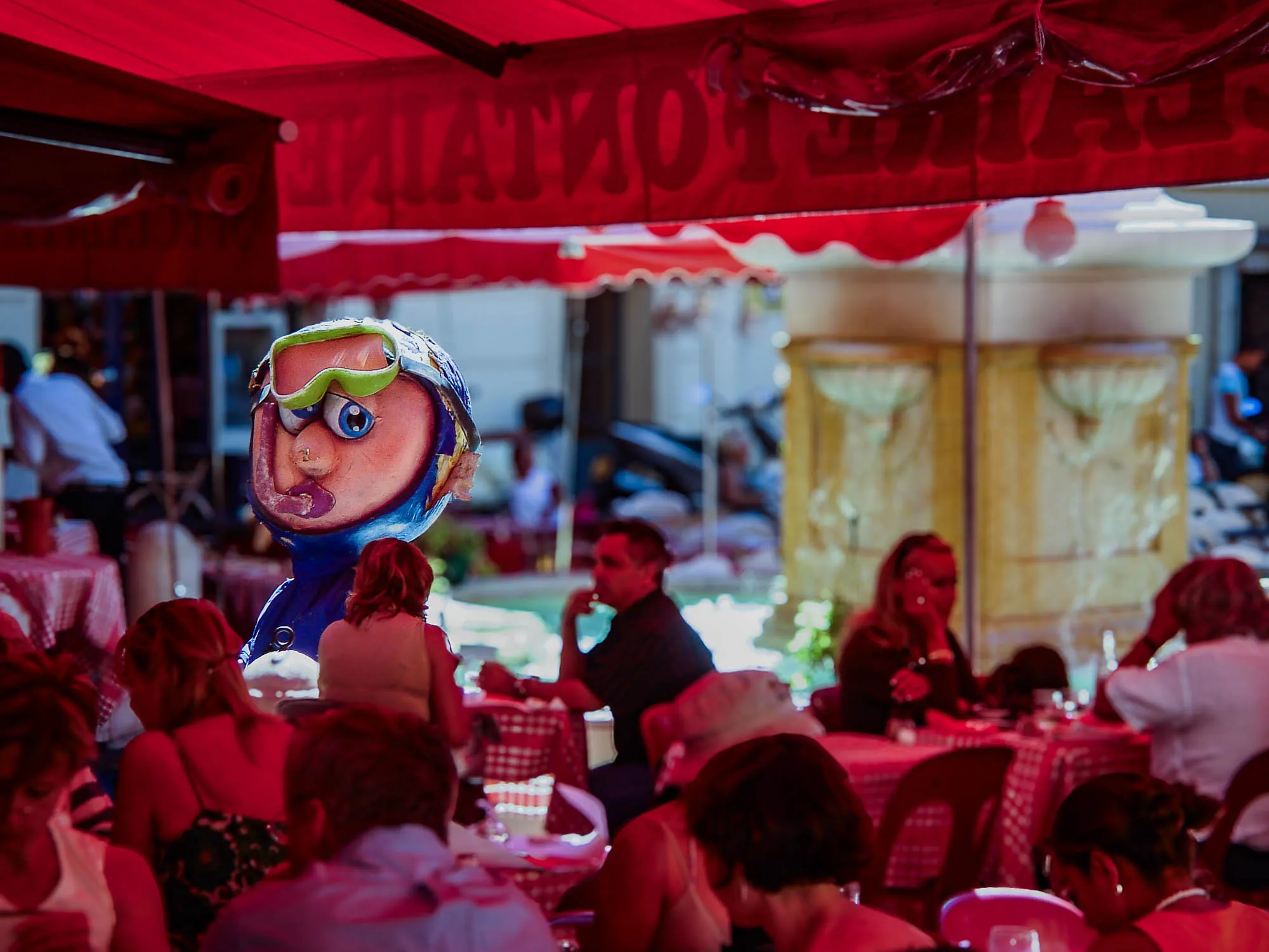 A seemingly papier-mâché cartoony scuba diver with a pink snorkel in his mouth, sideways blue eyes, and a pair of askew green snorkling goggles in a blue wetsuite that covers his head and body, stares across the outdoor dining tables of a restaruant. The patrons are under a red awning, giving them all a red glow. A fountain is in the background.