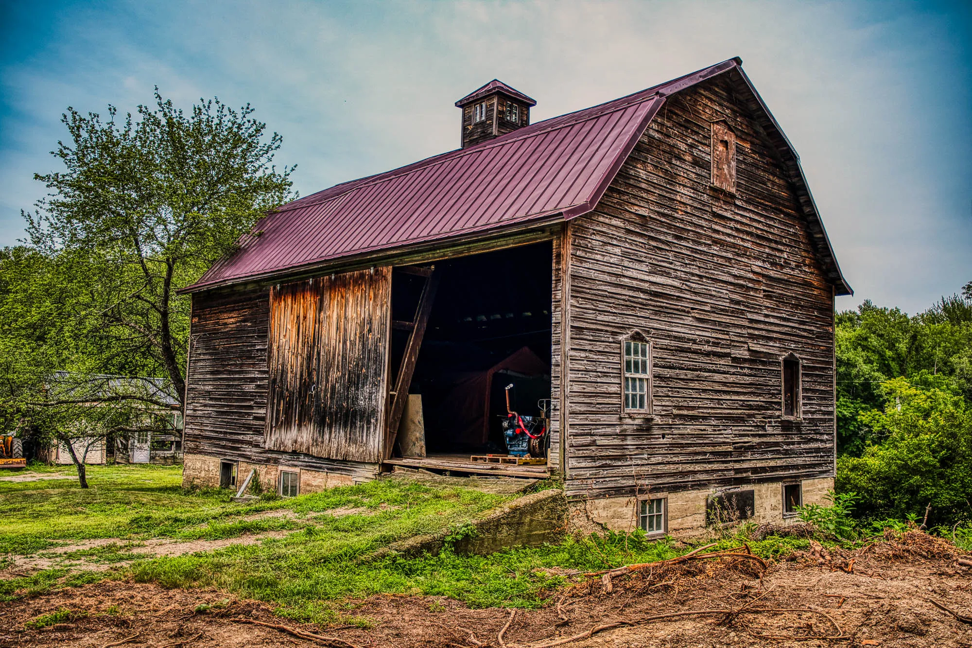 A wooden barn with the door open, a maroon roof with a roof lantern, and a grassy concrete ramp heading up into it