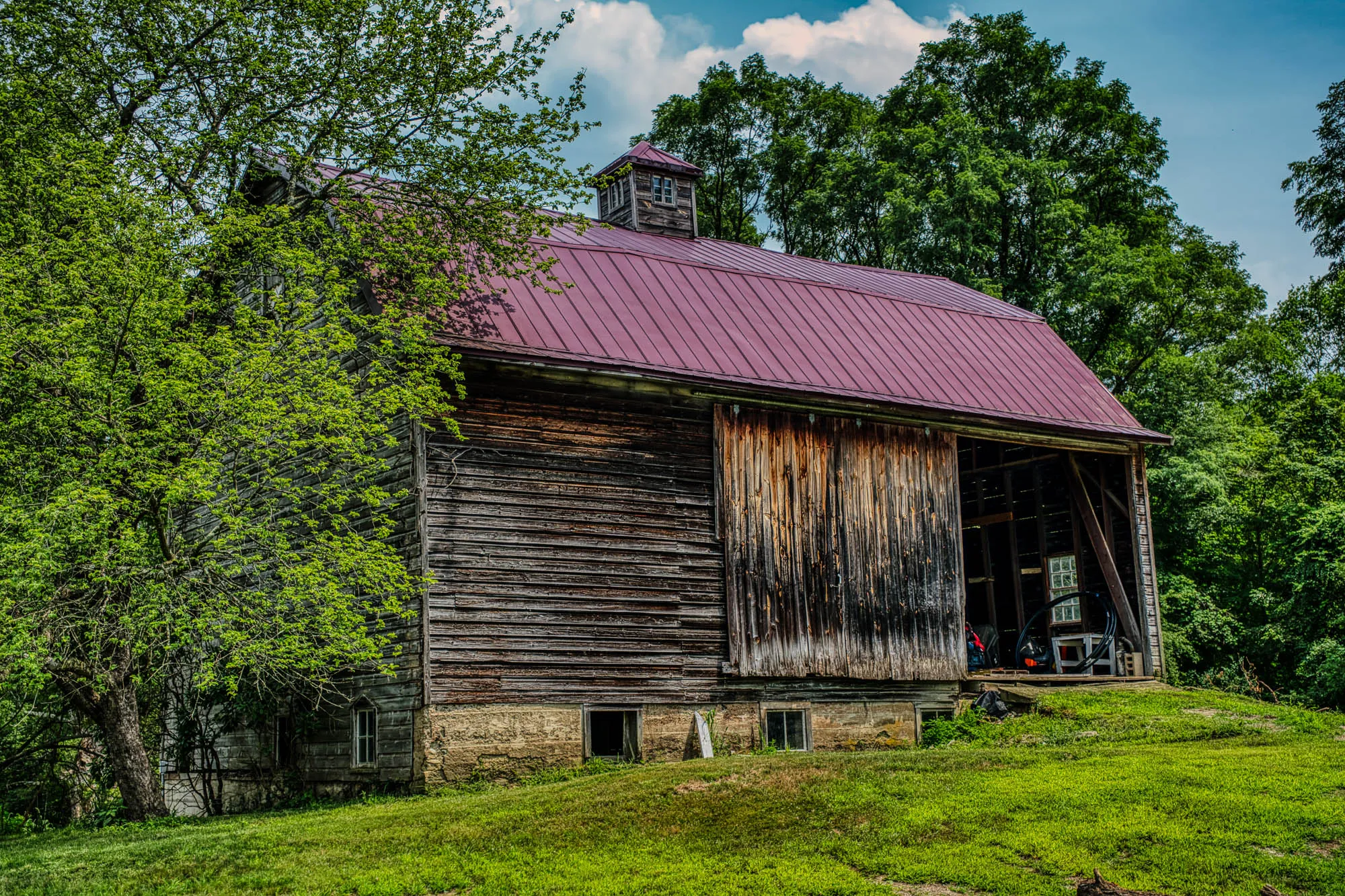 A wooden barn with the door open, a maroon roof with a roof lantern, and a grassy concrete ramp heading up into it. A tree is to its right.