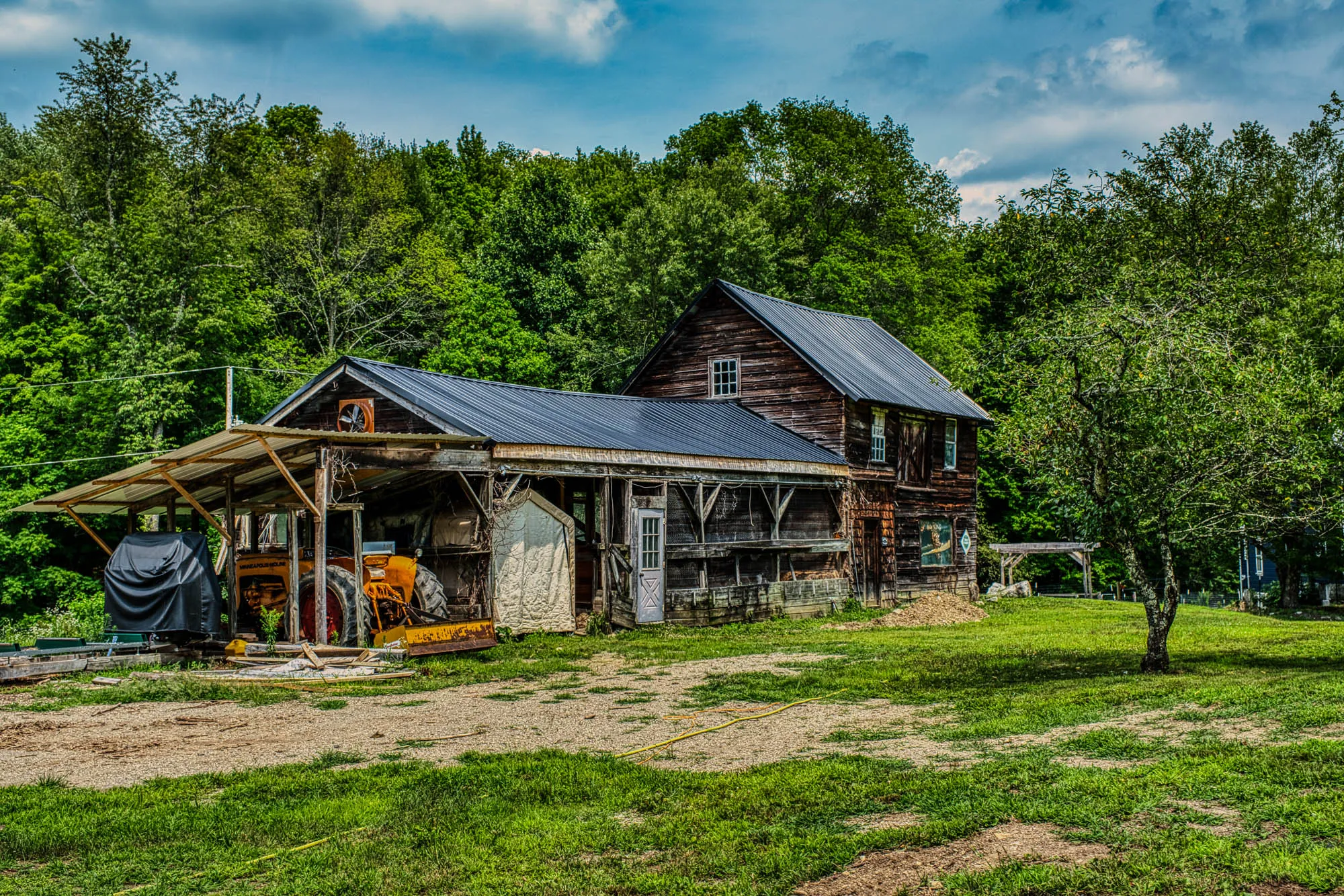 A two story wooden barn with windows and a stel roof, connected to a long enclused chicken run with front walls made by chicken wire, and a yellow tractor at the end. The grass in front is patchy, there's a tree in front, and there are trees behind it.