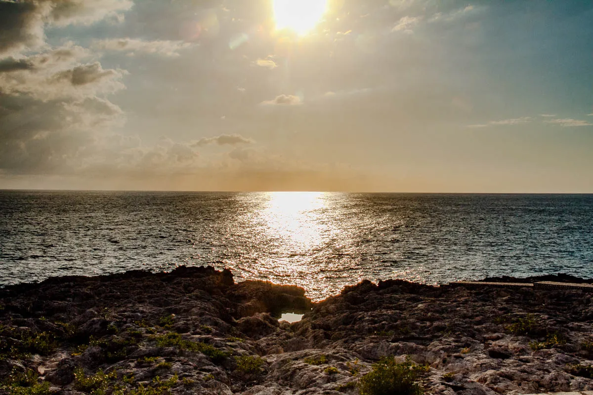 The image shows a sunset over the ocean. The sun is a bright white circle in the center of the image, shining through a few white clouds. The sky is a pale yellow, fading to blue at the edges of the image. The ocean is a deep blue, reflecting the sun's light in a path of shimmering gold.  In the foreground, the bottom of the image, there is a rocky outcrop. There is a small hole in the rock with a dark pool of water in it. The image gives the feeling of peaceful beauty and tranquility.