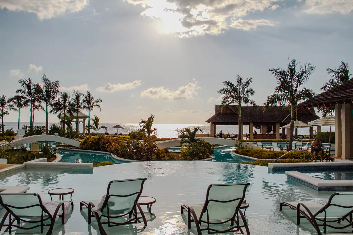 The image shows a beautiful resort scene. In the foreground, a large, rectangular pool extends outwards from the viewer. There are four lounge chairs arranged around the pool's perimeter, two on each side. The water is a refreshing blue and the pool is surrounded by lush greenery.  Beyond the pool, there is a winding path, a smaller, circular pool, and a white gazebo. In the distance, the ocean shimmers under a bright sun with white clouds floating above. To the right, there are two gazebos with wooden roofs, one behind the other. The gazebos have tables and chairs under them.  The scene is serene and inviting, with a tropical feel. There are palm trees throughout the area and a person sitting on a white lounge chair in the distance.  The sun is bright and shines through the clouds. 
