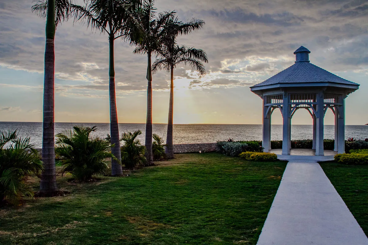 The image shows a white gazebo, sitting on a green lawn, with a stone wall behind it. There is a white walkway leading to the gazebo. The gazebo is on a cliff overlooking the ocean. A bright sun shines in the distance, and there are four palm trees in the foreground.