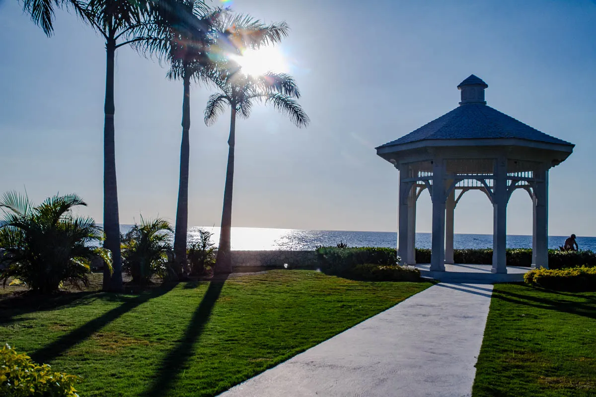 The image shows a white gazebo with a gray roof on a grassy beach, facing a calm, blue ocean with the sun shining in the background. The gazebo has a walkway leading up to it and is surrounded by lush green grass. There are three palm trees in the foreground, and the long shadows they cast suggest it is mid-day. On the right edge of the image, there's a person on a bicycle in the distance, also on the beach. The image gives off a peaceful, tropical vibe. 
