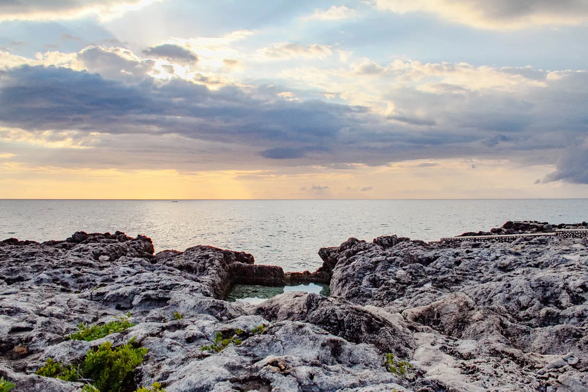 The image shows a rocky coastline with a small pool of water in the foreground. The rocks are a light grey color and appear rough and weathered. In the background, there is a vast expanse of blue ocean that stretches out to the horizon. The sky above is a mix of grey and white clouds, with some light breaking through in the center. The image appears to have been taken at sunset.  The sun is just below the horizon, and the sky is a soft pastel pink and yellow.  There are faint outlines of the shapes of small, distant islands on the horizon.