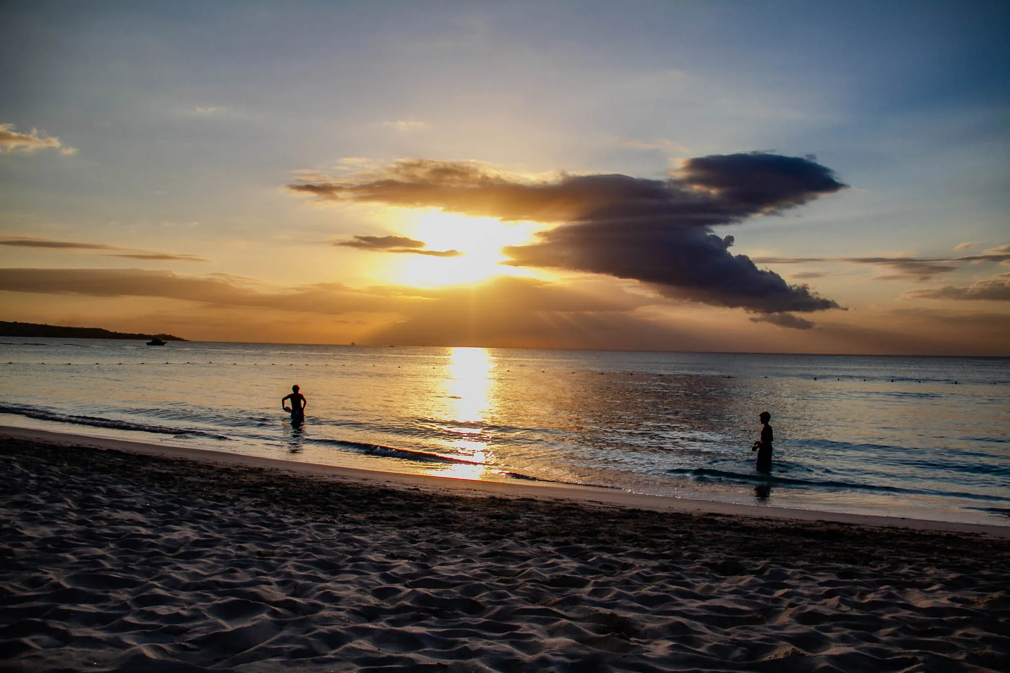 The image shows a sunset over a beach. The sun is setting behind a large dark cloud in the sky and is shining its light onto the ocean water. The ocean water is calm and reflects the light from the sun. There is a small boats in the distance and two people standing in the water near the shore. The sand on the beach is white and the sky is a light blue with hints of orange and yellow.