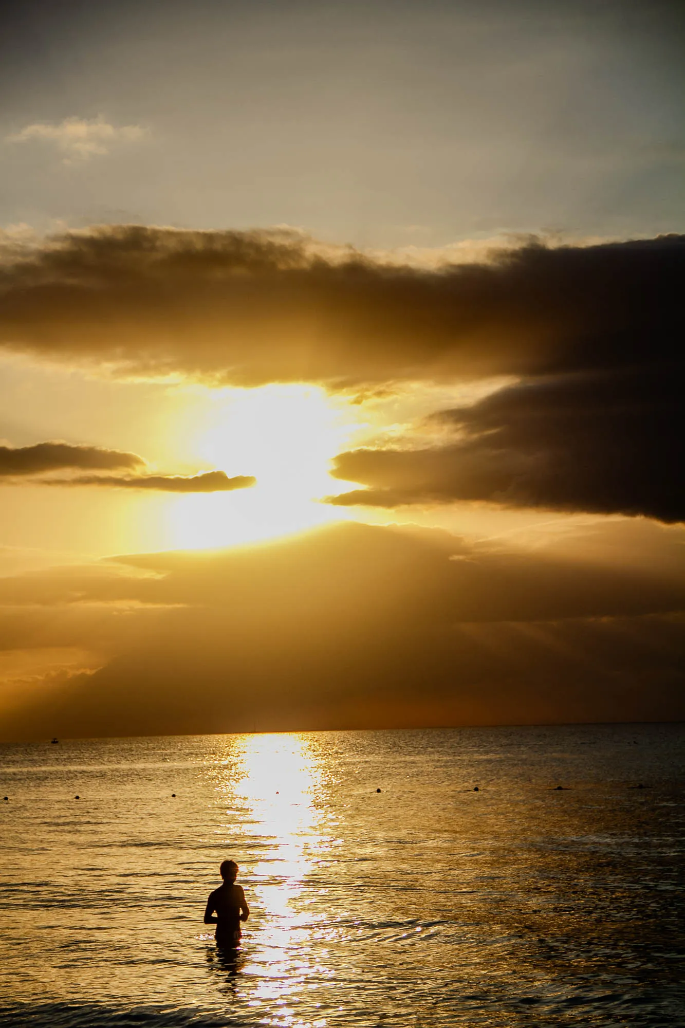 A silhouette of a person standing in the water, waist deep. The sun is setting in the background, casting a golden glow on the water and sky. There are clouds in the sky, some dark and some light. The water is calm and reflecting the golden light.