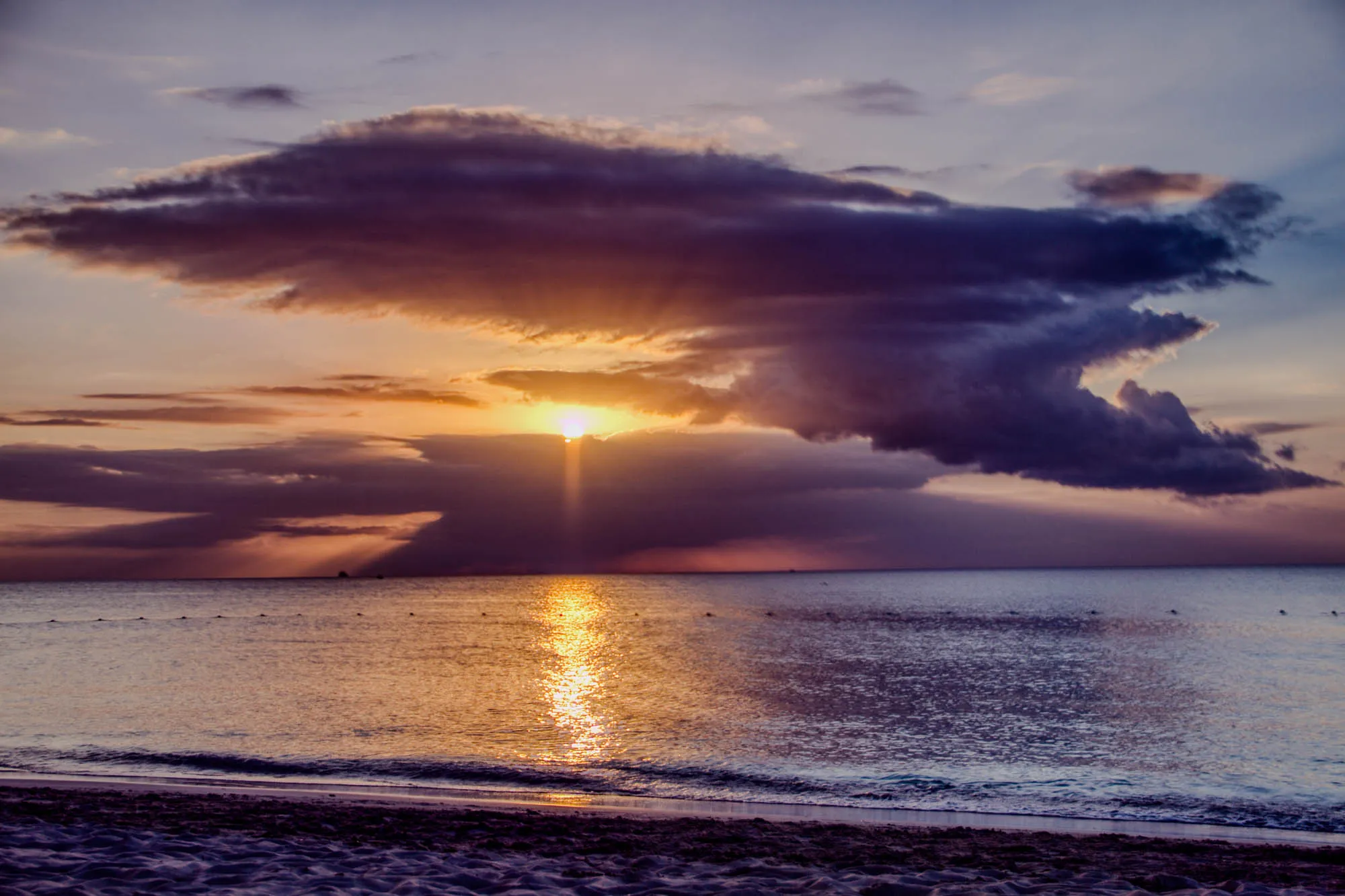 The image is a beautiful sunset over a calm ocean. The sky is a mixture of purple, pink, and orange with a large dark cloud covering the sun in the middle of the image. A bright beam of light from the sun shines down through the cloud and out across the water. The ocean is a calm blue with some light ripples and a sandy beach below. There is a slight wave at the edge of the shore.