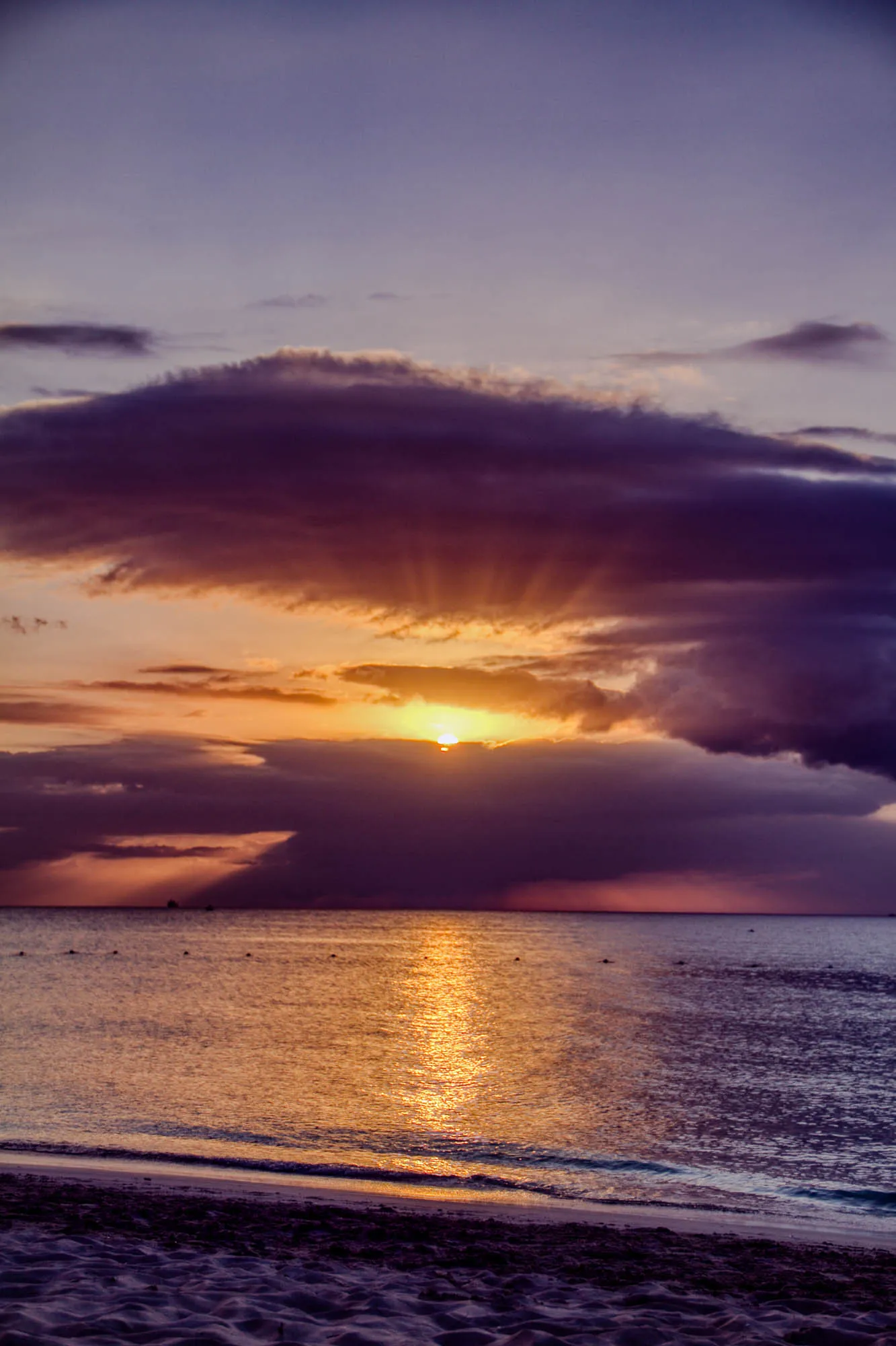 A sunset over a calm ocean with a sandy beach in the foreground. The sun is setting in the distance behind a large dark cloud with wispy edges. The sun is a bright yellow with rays of light shining through the clouds. The sky is a vibrant purple with shades of pink and orange. The ocean is a deep blue with sparkling reflections of the sun. The beach is a light brown color with a rippled texture.