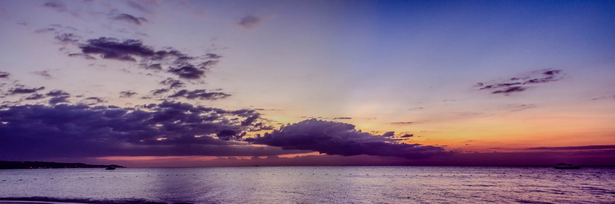 A wide shot of a beach at sunset. The sky is a mix of purple, blue, orange, and pink, with a large cloud in the center. The water is calm and a dark purple. A small silhouetted island can be seen in the distance.  