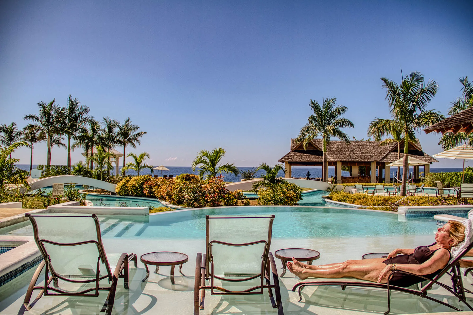 The image shows a woman lounging in a chair by a pool, with her legs stretched out in front of her. She is wearing a black swimsuit.  The pool is surrounded by palm trees and there is a small cabana behind her. The scene is very relaxing and inviting, with a clear blue sky and a calm ocean in the distance. There are two other chairs around the pool, both facing the ocean.  A small side table sits near the woman's chair. The pool is shaped like a crescent, with a shallow area near the edge.  There are some steps that lead down into the pool. The pool is empty except for the woman. The scene is very bright and sunny.