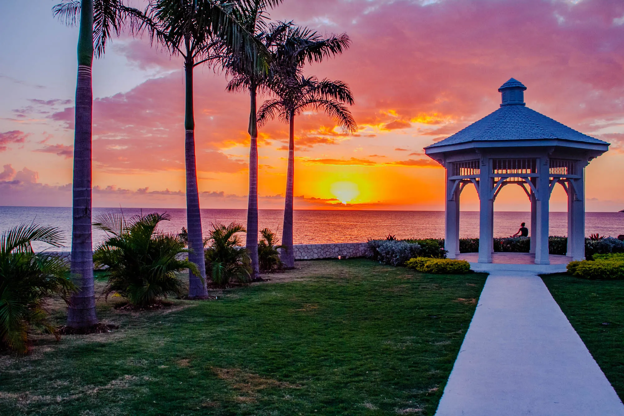 The image shows a beautiful sunset over the ocean. There are four palm trees in the foreground, and a white gazebo on a grassy hill. The sun is setting over the horizon, and the sky is a mix of pink, orange, and purple. The gazebo is empty except for a person sitting in the shade. There is a white path leading up to the gazebo. The sea is a dark blue, and the sky is clear.  The overall scene is serene and peaceful. 
