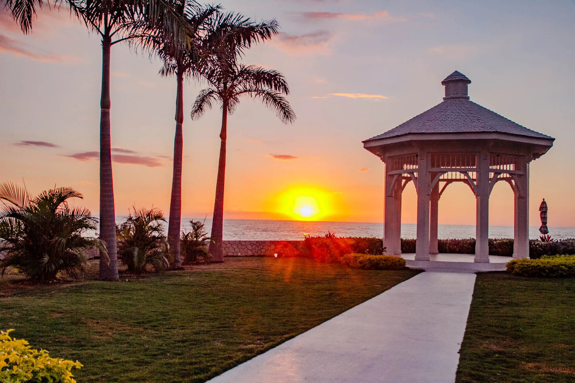 The image depicts a white gazebo standing on a grassy lawn with a paved path leading to it. The gazebo has a roof with a pointed top and open sides. Behind the gazebo is a calm body of water and a vivid orange sunset with a bright, round sun in the middle of the sky. In the foreground of the image, on the left side, are three tall palm trees. The scene suggests a tropical resort or beach location.