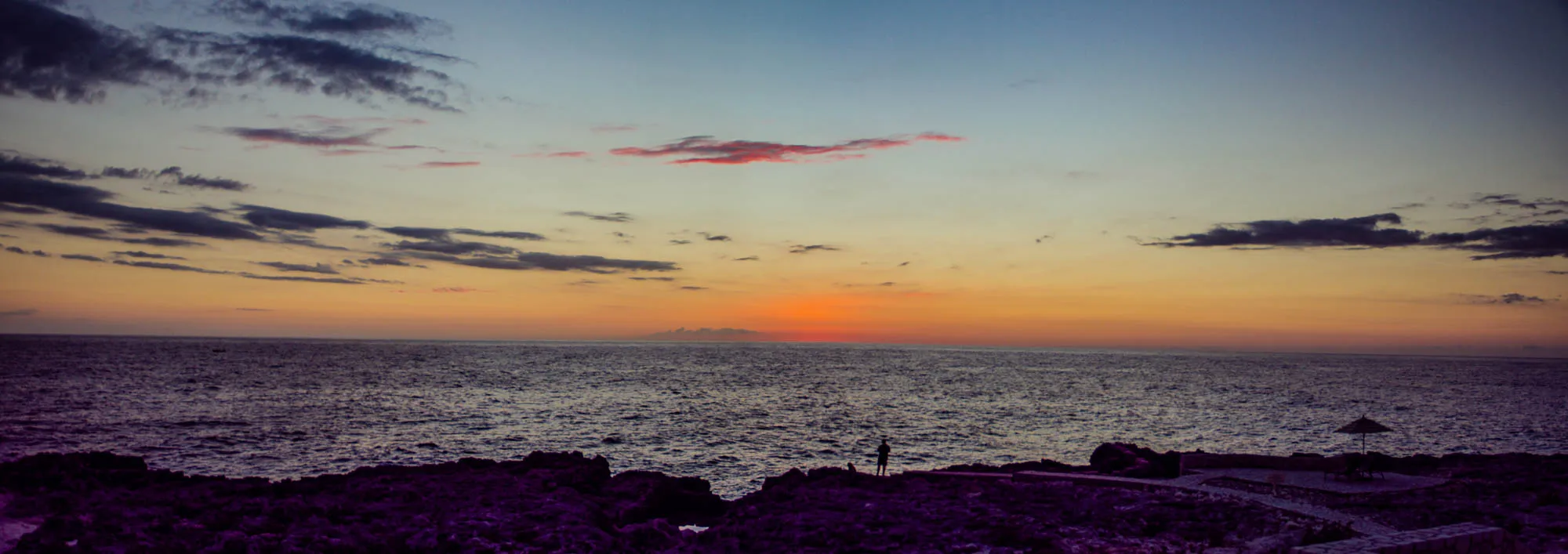 The image shows a sunset over a calm, dark blue ocean. The sky is a mix of pink, orange, and blue, with wispy clouds and a single dark cloud near the horizon. In the foreground, there's a dark rocky shore with a single person standing on it. The person is silhouetted against the orange sky, and appears to be wearing a light-colored shirt. To the right of the person, there's a small, dark umbrella on a beach.  The image is taken at a wide angle, with the horizon appearing in the middle of the frame. It conveys a sense of tranquility and peace. 
