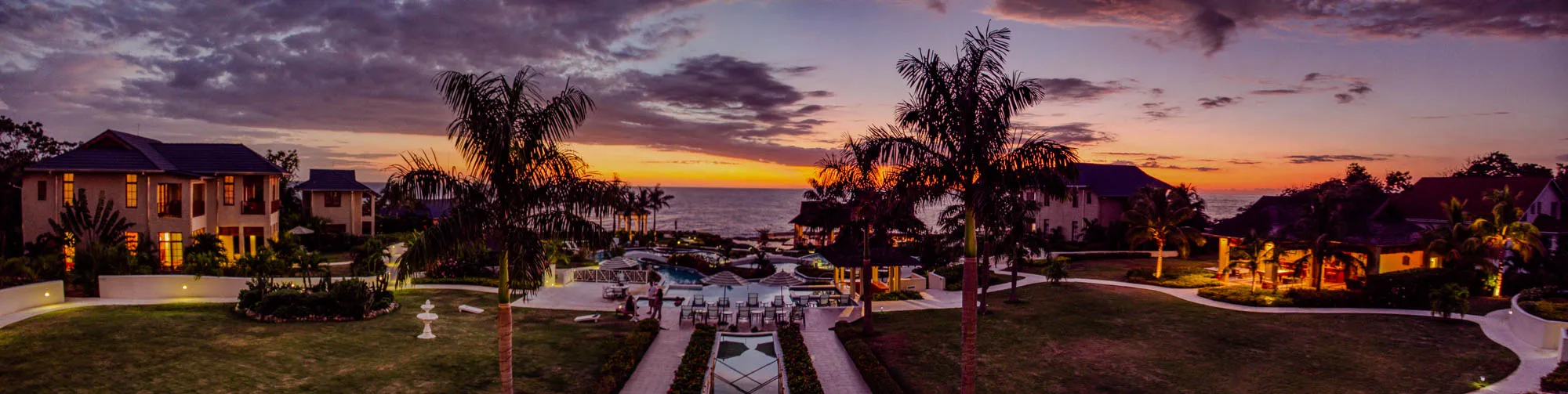 The image shows a beautiful resort at sunset. There are several buildings in the foreground, some with lit windows, and a large swimming pool with white chairs around it. In the background, there is a calm ocean with a vibrant sunset behind it.  There are many palm trees throughout the scene, some of which are silhouetted against the sunset. The sky is a mix of purple, pink, and orange, creating a picturesque scene. The image is captured from a slightly elevated perspective, giving a panoramic view of the resort. The lighting is warm and inviting, creating a sense of relaxation and tranquility. 
