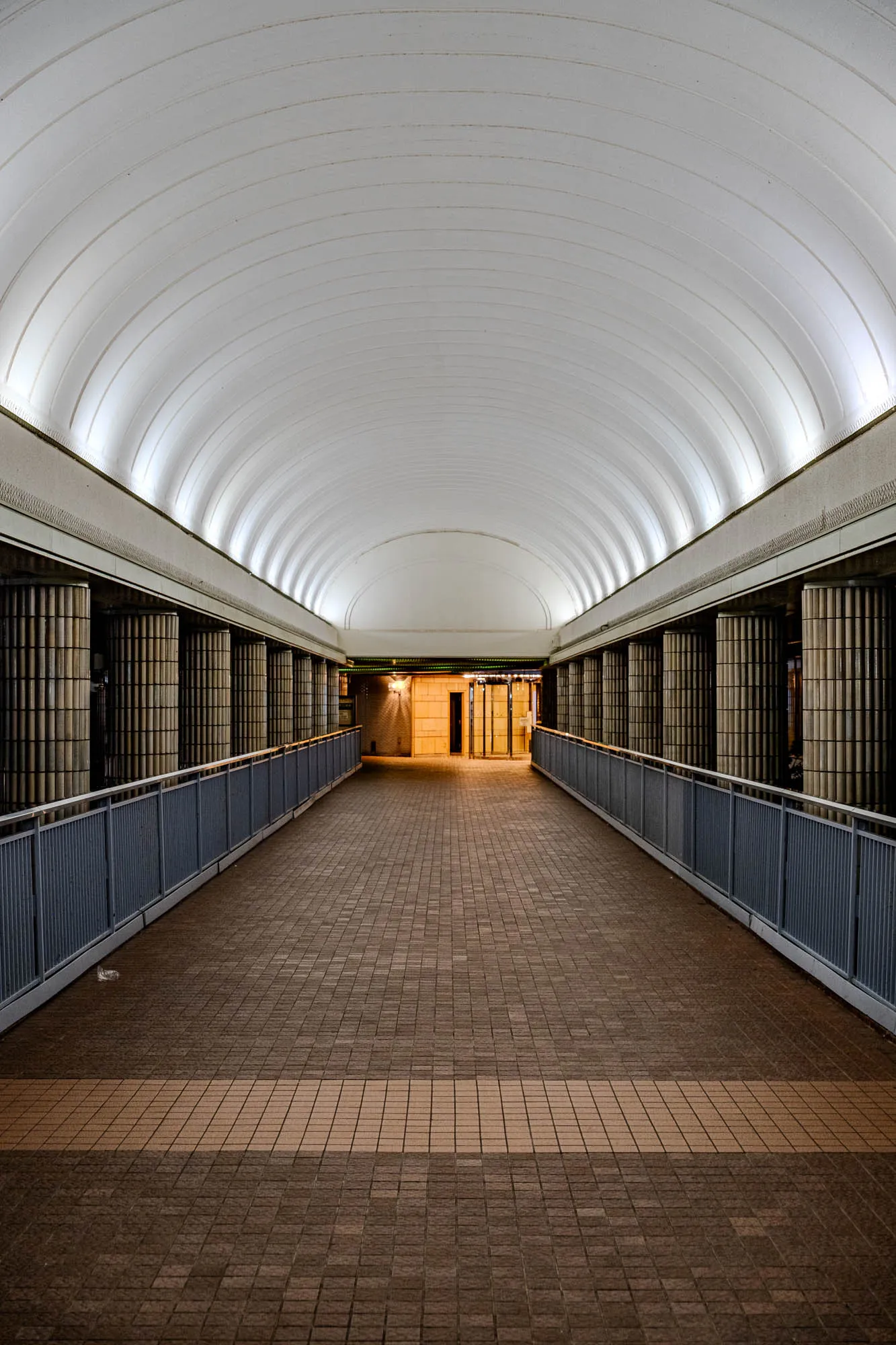 A symmetrical, one-point perspective view looks down a long, covered pedestrian bridge defined by a soaring, white barrel-vaulted ceiling with illuminated structural ribs. Flanking the central walkway are rows of cylindrical columns clad in glossy beige tiles, separated from the path by industrial blue-grey metal railings. The floor is paved with small, earth-toned square tiles that create textured leading lines toward a distant, warm-lit entrance at the far end of the corridor. The wide-angle composition emphasizes the depth and geometric repetition of the architecture.