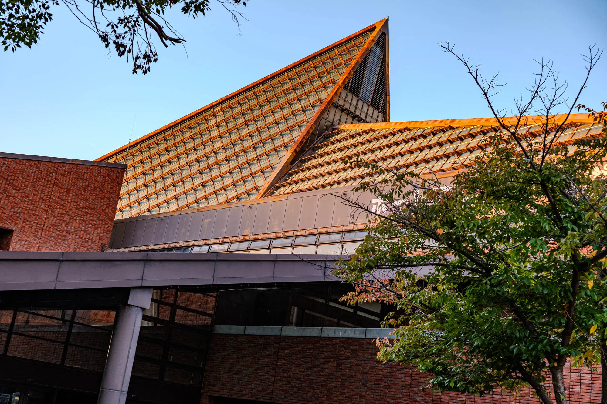 A modern architectural structure is dominated by a sharp, triangular roof peak clad in a tessellated grid of copper-toned scales that shimmer under warm, directional light. A block of reddish-brown brick anchors the left side of the building, contrasting with the geometric intricacy of the angled roofline. Below the peak, a flat, grey overhang extends horizontally, supported by a sturdy column near the darkened glass windows at the base. The right foreground is framed by the green and turning leaves of a tree, adding organic texture to the scene's rigid, angular composition against a clear blue sky.