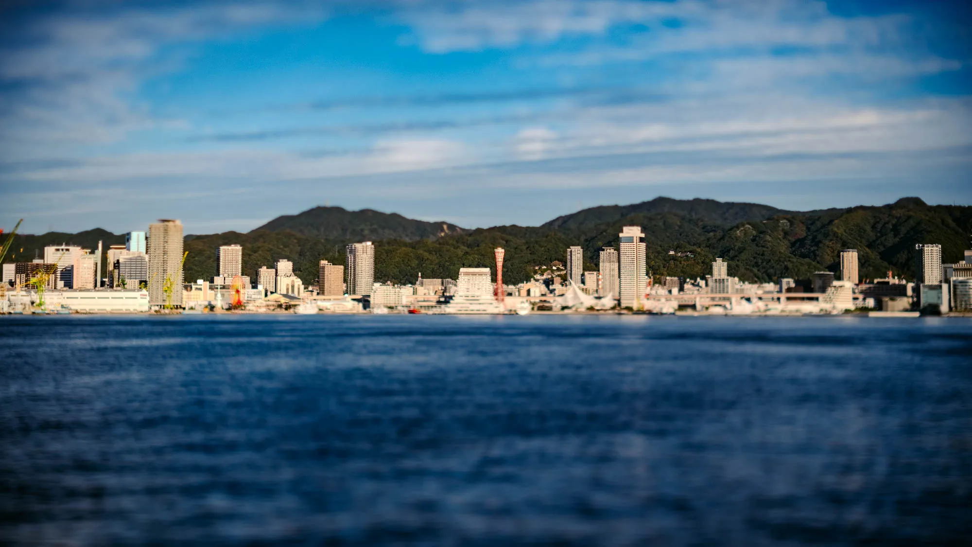 A coastal city skyline, rendered with selective focus, stretches across the midground, appearing like a miniature model. Tall, light-colored high-rise buildings and other structures line the water's edge, set against a backdrop of dark green, densely forested mountains. The deep blue water in the foreground and the sky above the mountains are softly blurred, characteristic of a tilt-shift effect. A striking red tower rises prominently from the city, beneath a bright blue sky dotted with wispy white clouds.