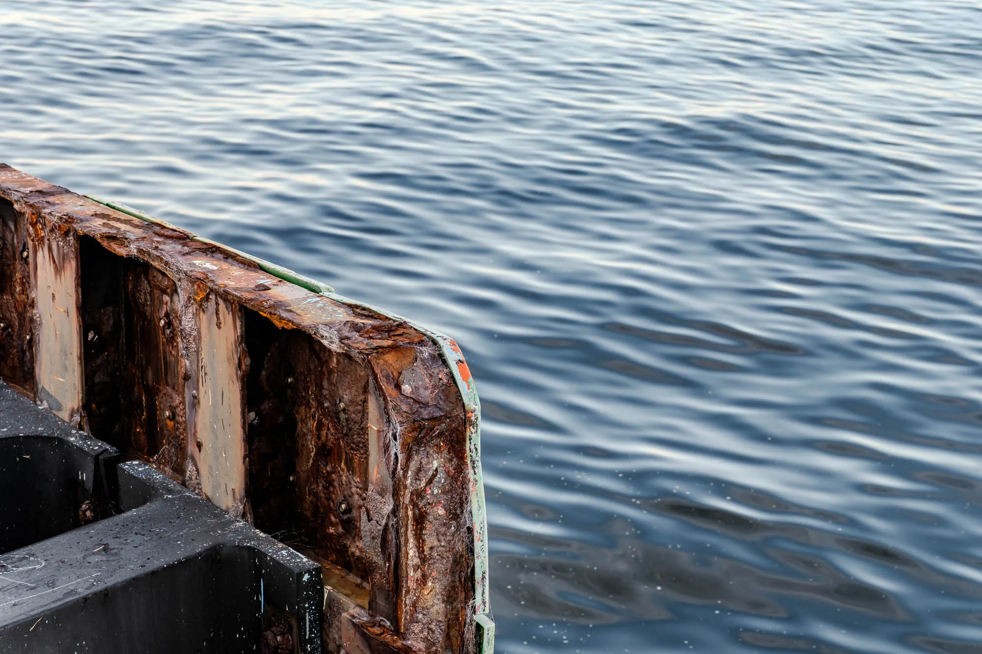 A heavily corroded metal barrier dominates the left foreground with thick layers of flaking, vibrant orange and deep brown rust. Its rigid, decaying vertical panels contrast with a smooth, black industrial bumper attached at the bottom edge. To the right, the dark blue water fills the background, textured by soft, rhythmic ripples that reflect the ambient light. The composition emphasizes the tactile difference between the jagged, oxidized metal and the fluid, undulating surface of the sea.