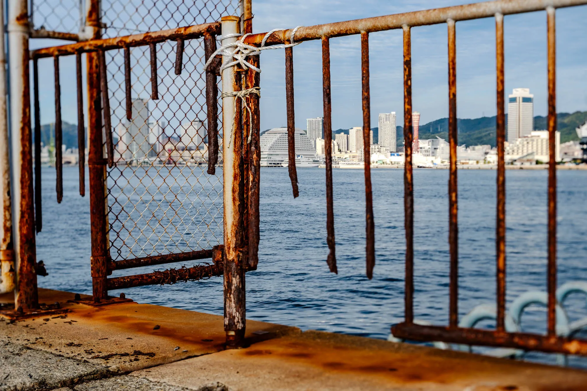 A heavily rusted iron fence stands in sharp focus against a sunlit harbor, its vertical bars deeply corroded with bright orange oxidation and several detached struts hanging loose near the concrete base. A section of chain-link mesh is lashed to the railing with white rope, emphasizing the decaying industrial texture of the foreground. Across the blue water, the cityscape of Kobe is rendered soft by a shallow depth of field, though the red lattice structure of the Port Tower and the white, curved silhouette of the Meriken Park Oriental Hotel remain identifiable. The composition concludes with the faint, hazy outline of the Rokko Mountains rising behind the urban skyline.