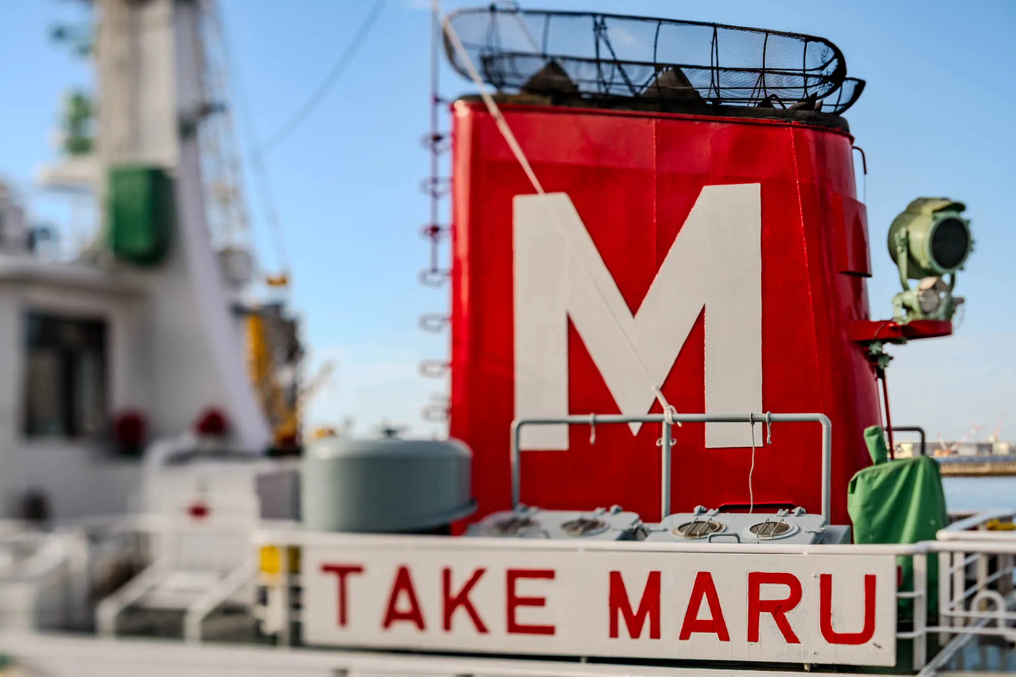 A bright red ship's funnel anchors the composition, featuring a prominent white capital "M" painted on its side. Below this structure, a white sign reads "TAKE MARU" in red block letters, positioned near a green navigational device. The background elements, including the ship's white masts and the blue sky, are softened into a heavy blur by a tilt-shift effect.