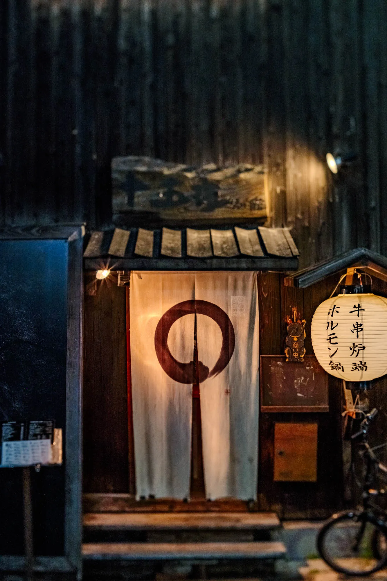 Warm artificial light emanates from a traditional Japanese entryway, contrasting with the dark, vertical wooden siding of the building's facade. White fabric curtains hang centrally, adorned with a large, rustic reddish-brown circle painted in a brushstroke style. To the right, a glowing paper lantern with vertical Japanese text hangs above a small wooden lucky cat figurine attached to the wall. A tilt-shift photographic technique blurs the upper wooden textures and the bicycle wheel in the lower foreground, keeping the focus sharply on the illuminated entrance.