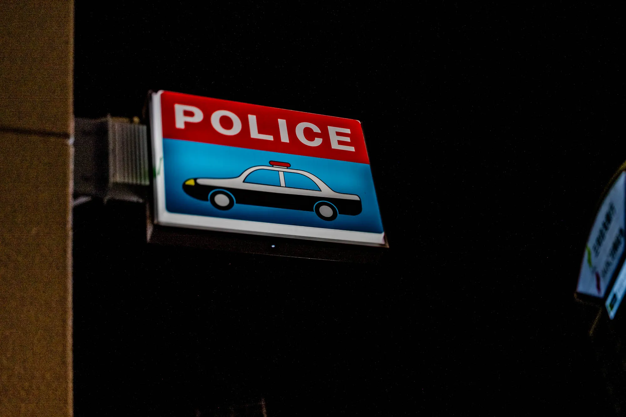 An illuminated rectangular sign projects from a textured, beige wall on the left, glowing intensely against the pitch-black void of a late-night setting. The box light features a red upper band displaying the word "POLICE" in bold white capitals, while the blue section below illustrates a simplified, cartoon-like profile of a white patrol car with a red siren. Sharp focus renders the sign's vivid colors and clean lines distinct, contrasting with a blurry, partially visible element on the far right edge. The darkness surrounding the fixture isolates it completely, emphasizing its function as a beacon in the night.