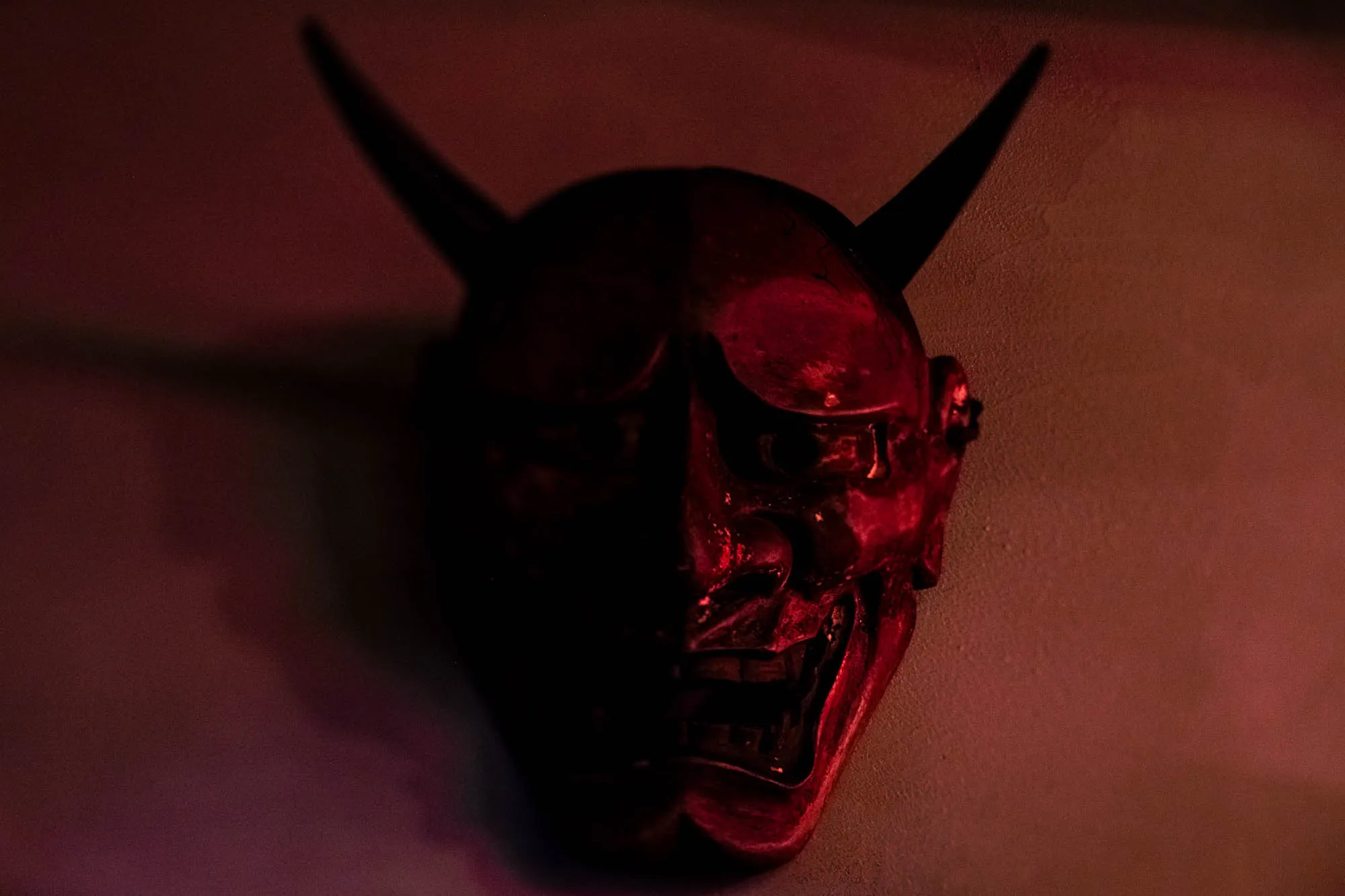 A traditional Japanese Hannya mask hangs on a textured wall, illuminated by a deep crimson light that casts the left half of the face into heavy shadow. Two sharp, curved horns rise from the forehead, framing staring eyes and a mouth fixed in a wide, toothy grimace. The dramatic, low-key lighting emphasizes the glossy, lacquered texture of the illuminated cheek and nose while obscuring the rest in darkness.