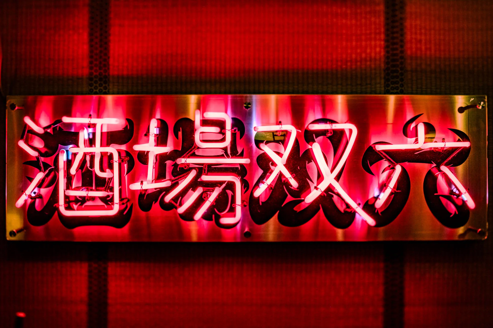A rectangular brushed metal sign displays four prominent Japanese kanji characters traced in bright pinkish-white neon tubing. The glowing tubes float above corresponding black painted strokes on the plate, creating a bold, high-contrast calligraphic design. This intense illumination casts a saturated red wash over the metallic surface and the dark, mesh-textured wall visible in the background.