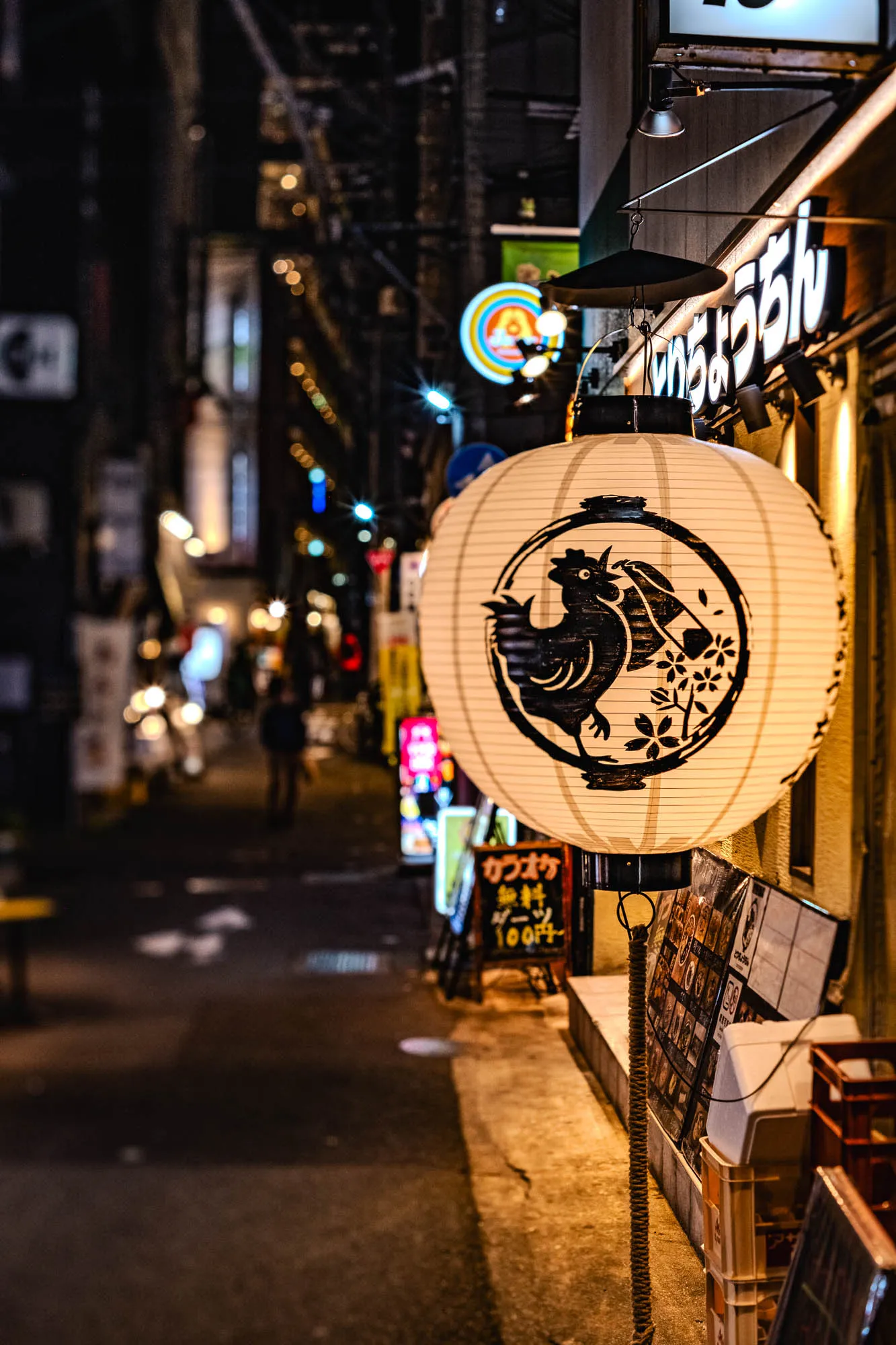 A luminous white paper lantern dominates the right foreground, featuring a bold black ink illustration of a rooster encircled by floral motifs. It hangs outside a Japanese establishment, casting warm yellow light onto a chalkboard menu and plastic crates near the entrance. A shallow depth of field renders the adjacent narrow alleyway into a soft blur, turning distant streetlights and neon signs into hazy orbs of blue and amber. The dark, atmospheric street creates a high-contrast backdrop that isolates the intricate texture and glow of the lantern.