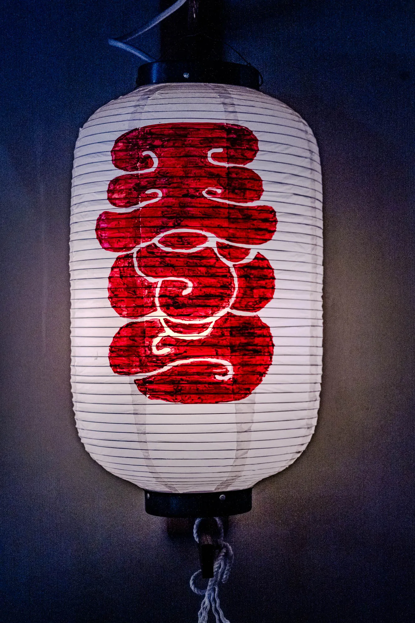 A cylindrical white paper lantern hangs vertically, illuminated from within to cast a warm glow against a dark, shadowed background. Rigid horizontal ribs structure the paper surface, which bears a large, stylized red character painted with thick, bold strokes. The internal light emphasizes the paper's texture and the high contrast between the vibrant red pigment and the pale body. A braided white cord dangles loosely from the lantern's black circular base.