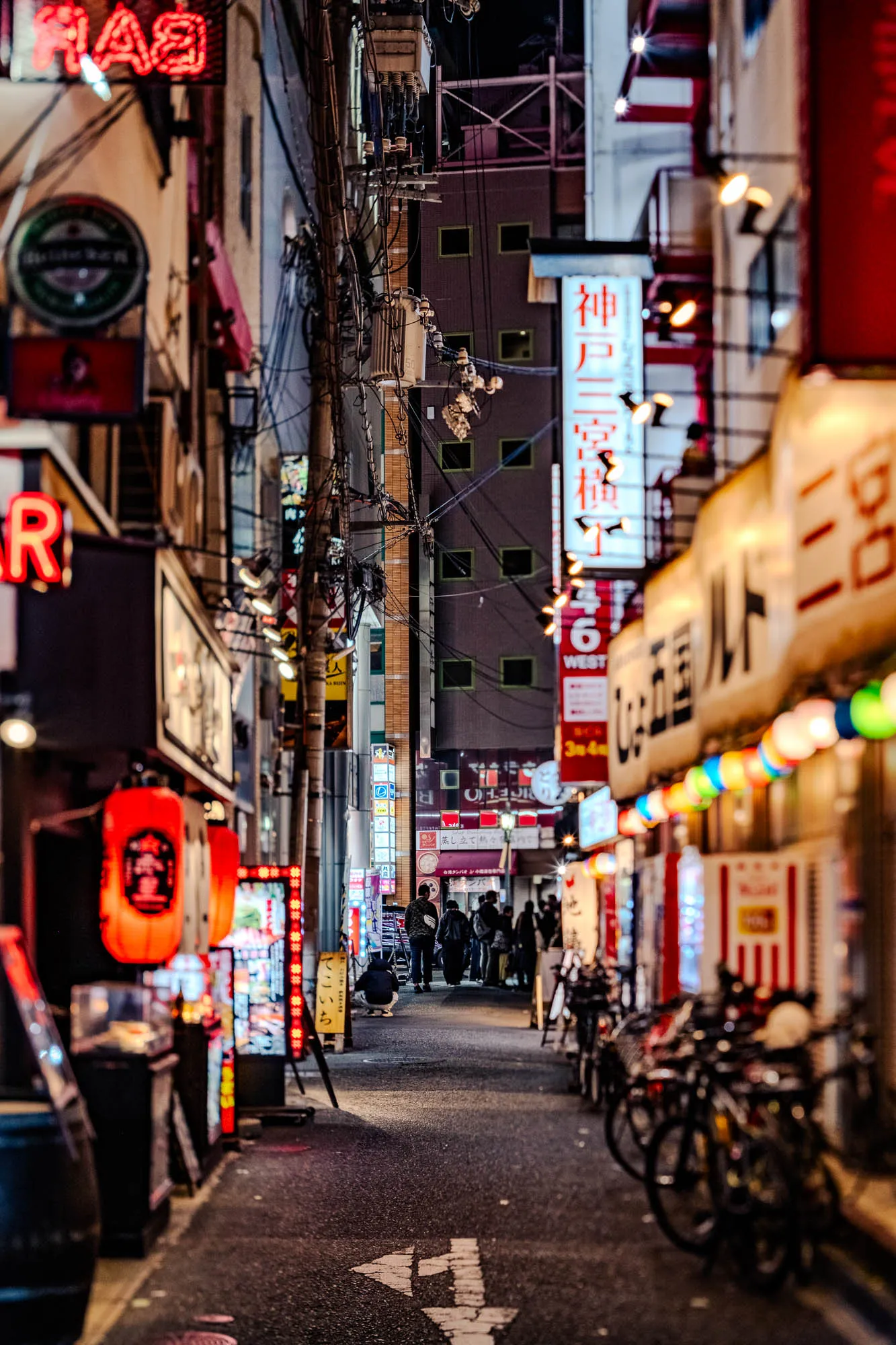 Vertical neon signs and glowing red paper lanterns illuminate a narrow, crowded alleyway in Kobe, Japan at night. A chaotic web of power lines and transformers hangs overhead, framing the corridor where pedestrians walk away into the distance. To the right, a row of parked bicycles renders softly out of focus due to a shallow depth of field, contrasting with the sharp details of the mid-ground signage. The asphalt street is marked with a white directional arrow, leading the eye deep into the vibrant urban scene.
