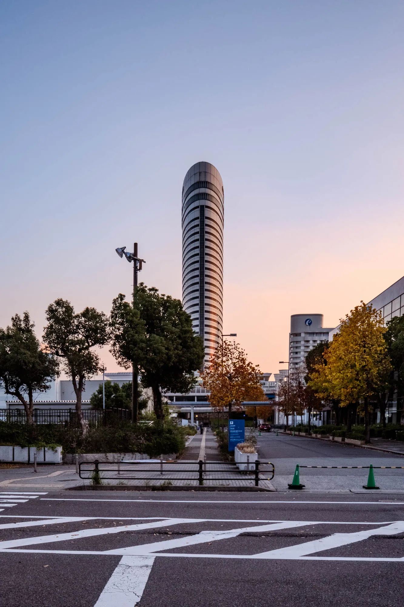 The oval, ribbed tower of the Kobe Portopia Hotel anchors the composition, rising centrally against a sky transitioning from cool cerulean to a soft dawn apricot. In the foreground, stark white road markings on asphalt create geometric leading lines that guide the gaze past a low black railing and toward a pathway flanked by trees displaying yellow and green autumn foliage. The early morning light casts the street level in cool shadows while illuminating the eastern curve of the skyscraper's white facade, highlighting its vertical symmetry. A quiet urban atmosphere pervades the scene, with traffic cones and a blue sign marking the entrance to the complex amidst the stillness of sunrise.