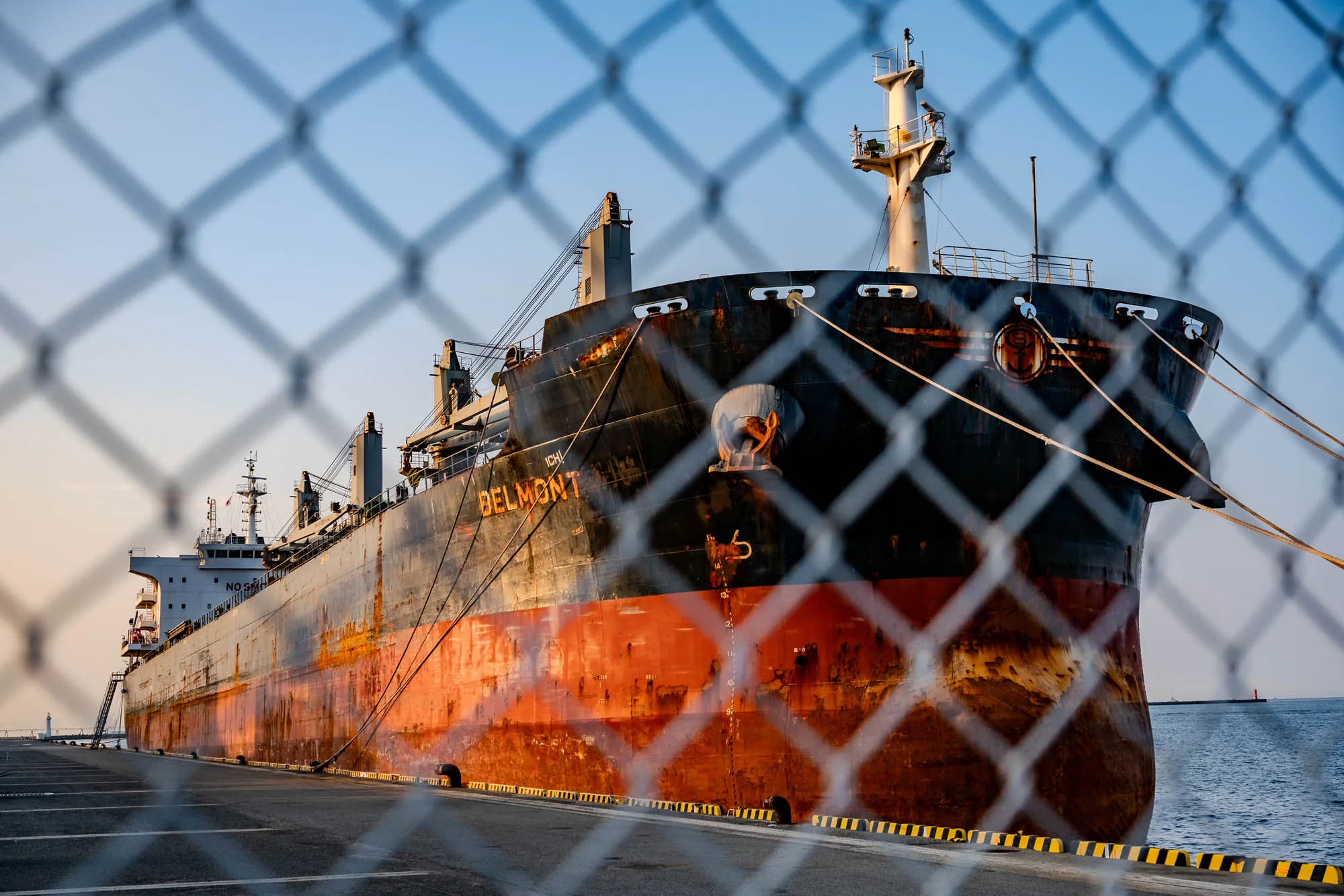 A massive cargo ship named "BELMONT" is docked at Port of Kobe, its hull defined by a weathered combination of black upper paint and heavily rusted orange anti-fouling paint below. The view is framed through a chain-link fence in the immediate foreground, which is rendered into soft, out-of-focus diamond shapes due to a shallow depth of field. Golden morning sunlight strikes the starboard side of the vessel, accentuating the industrial textures and the taut mooring lines connecting the ship to the concrete pier. To the right, the calm water of the harbor extends toward a clear blue sky, suggesting an early autumn morning.