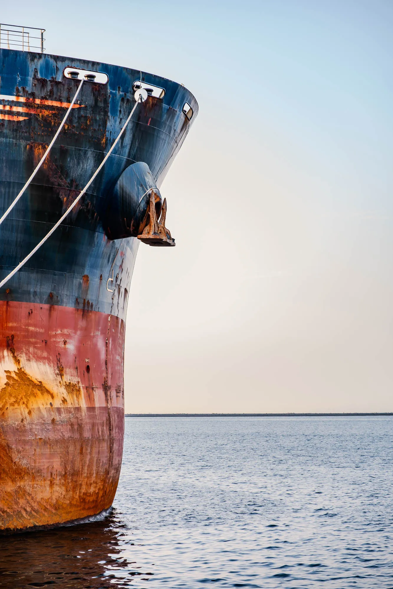 The towering bow of a massive cargo vessel dominates the left foreground, its hull painted in bands of deep navy and faded crimson that are heavily streaked with vibrant orange corrosion. Two taut white mooring lines cut diagonally across the weathered steel, leading from the upper deck railing past a heavy, rusted anchor resting in its hawsepipe. The ship sits in the calm, rippled waters of Osaka Bay, illuminated by soft morning light that emphasizes the texture of peeling paint against the smooth, pale gradient of the sky. To the right, the composition opens into vast negative space, broken only by a distant, flat breakwater on the horizon.