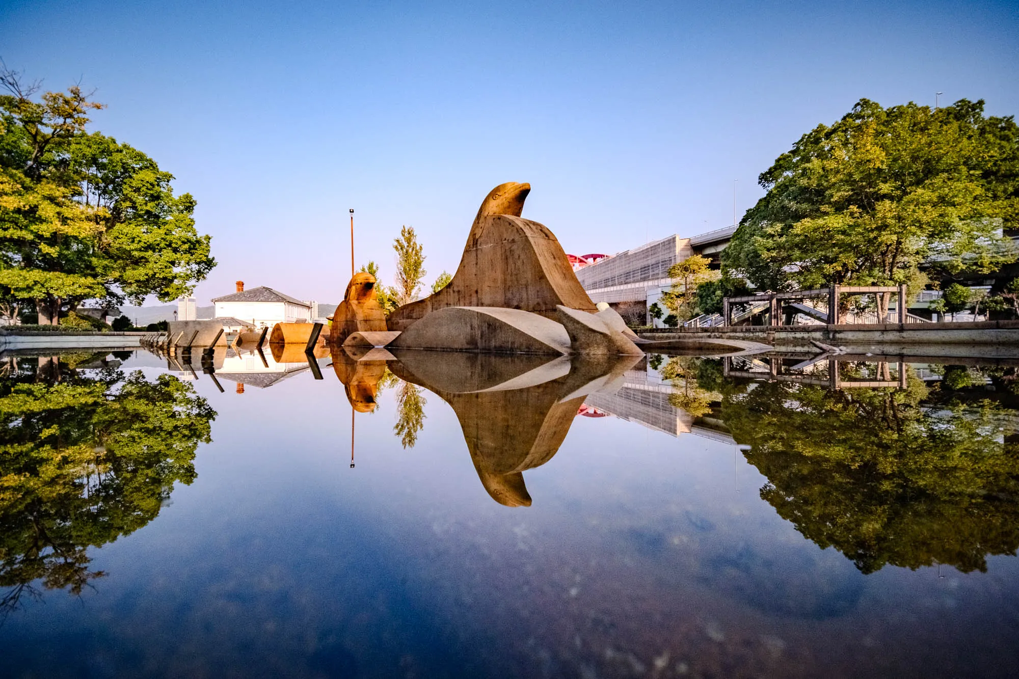 A tan, abstract sculpture resembling a stylized marine form anchors a tranquil reflecting pool within Kobe's Meriken Park. The geometric stone structure is mirrored perfectly in the still water below, creating a symmetrical composition flanked by vibrant green trees on either bank. In the background, the historic white facade of the Former Kobe Foreign Settlement 15th Building contrasts with the modern concrete span of the Hanshin Expressway. The scene is bathed in clear morning light, with a deep azure sky graduating to a soft horizon.